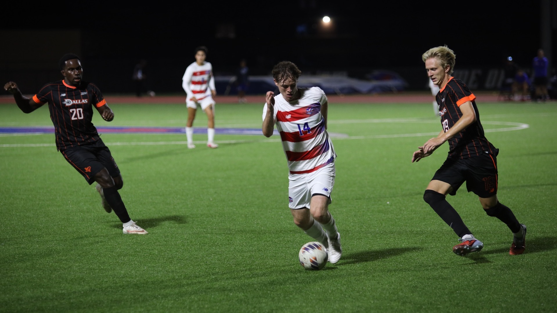 Lubbock Christian midfielder Tom Unwin (white jersey, #14) dribbles the soccer ball between two UT Dallas defenders (black jerseys) on a turf field at the LCU Soccer & Track Complex. 