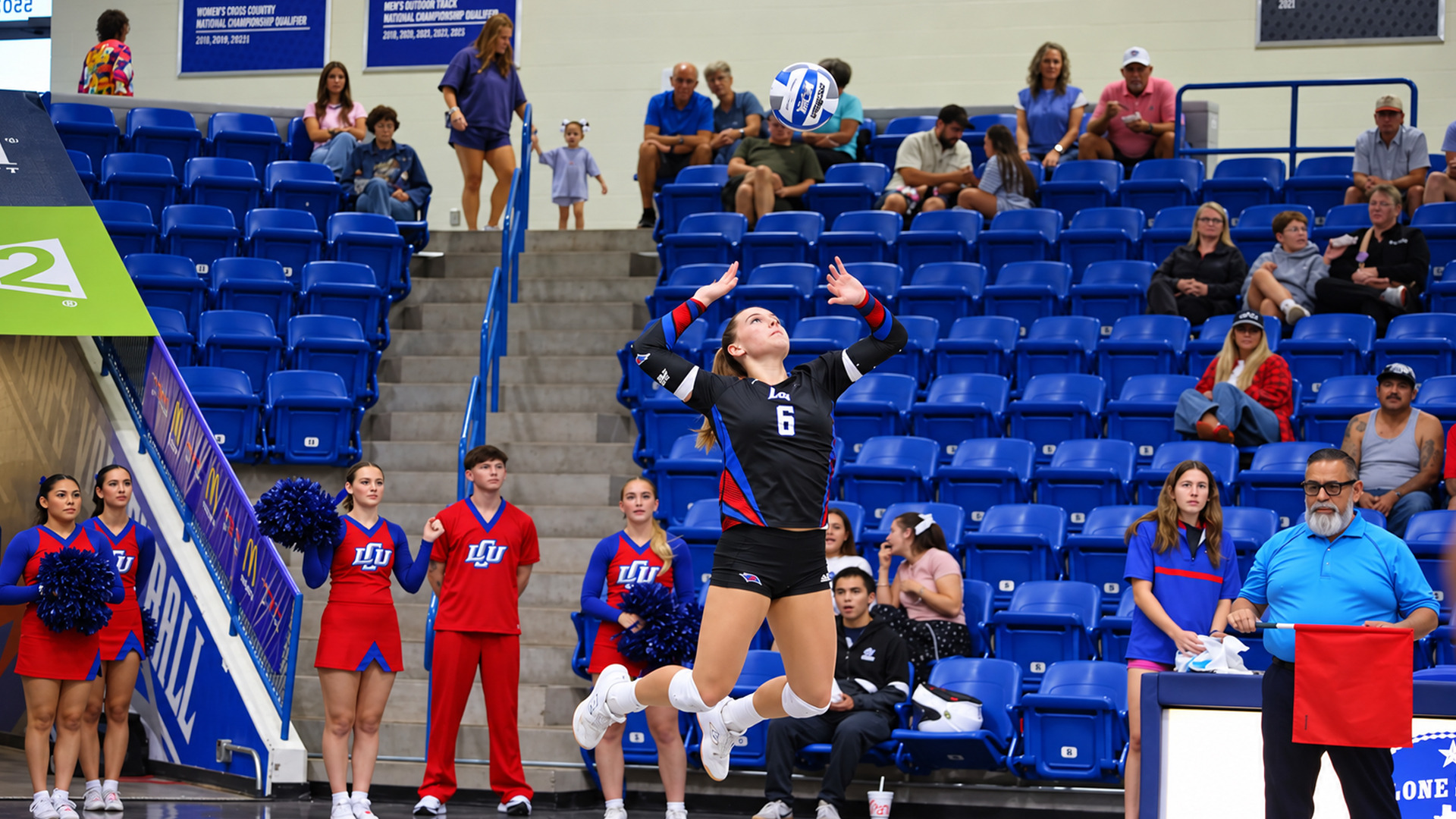 Action photo of LCU player #6, in a black jersey, leaping off the floor to serve the ball, both arms raised overhead as she tracks it. Cheerleaders in red and blue uniforms with pom-poms stand along the left, and a line judge in a light blue shirt holds a red flag at right. Blue stadium seating with spectators fills the background.
