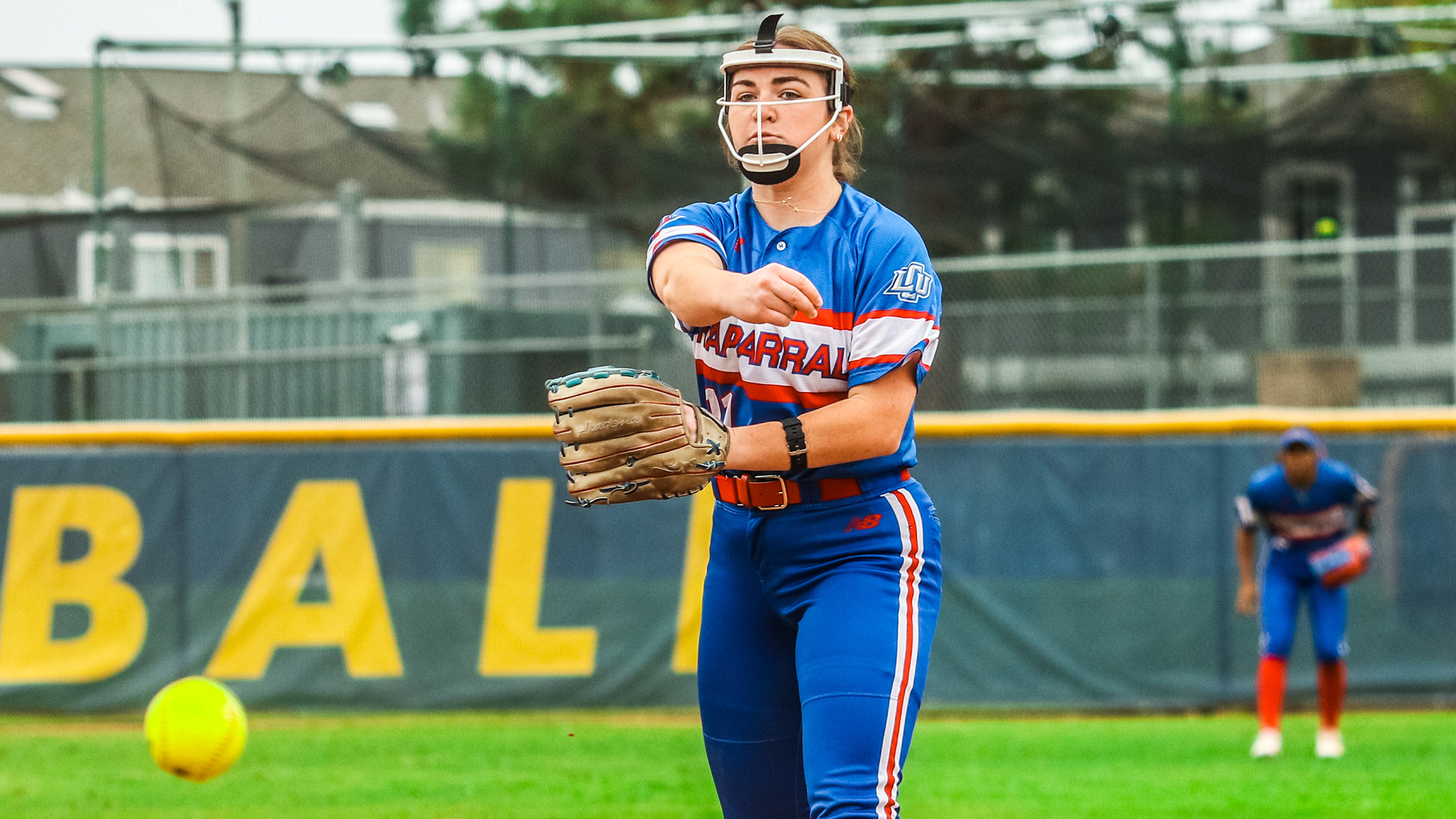 LCU softball pitcher in blue striped 'CHAPARRALS' uniform number 21 with white fielder's mask completing pitching delivery. Yellow softball visible in lower left mid-flight. Player wears tan glove and red belt. Green outfield wall with yellow 'BALL' lettering in background. Teammate in blue uniform visible in right background. Sunny day game action showing pitch release at Vanguard softball field