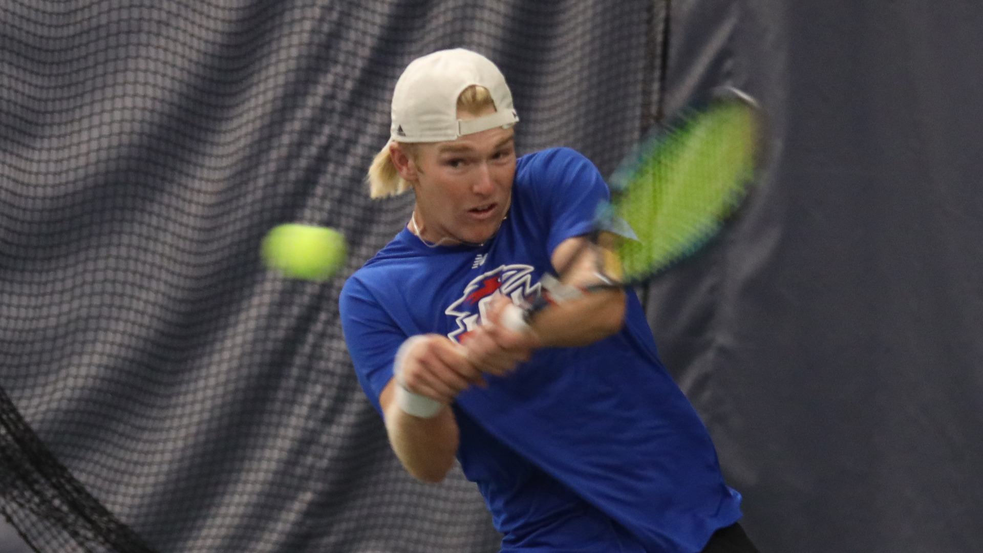 LCU men's tennis player in blue shirt and white backwards cap hitting two-handed backhand with green and black racket on indoor court. Yellow-green tennis ball visible in motion on left. Player focused on shot with gray windscreen background. Indoor tennis facility action shot showing dynamic stroke mechanics during ITA National Team Indoor Championships match against Washburn