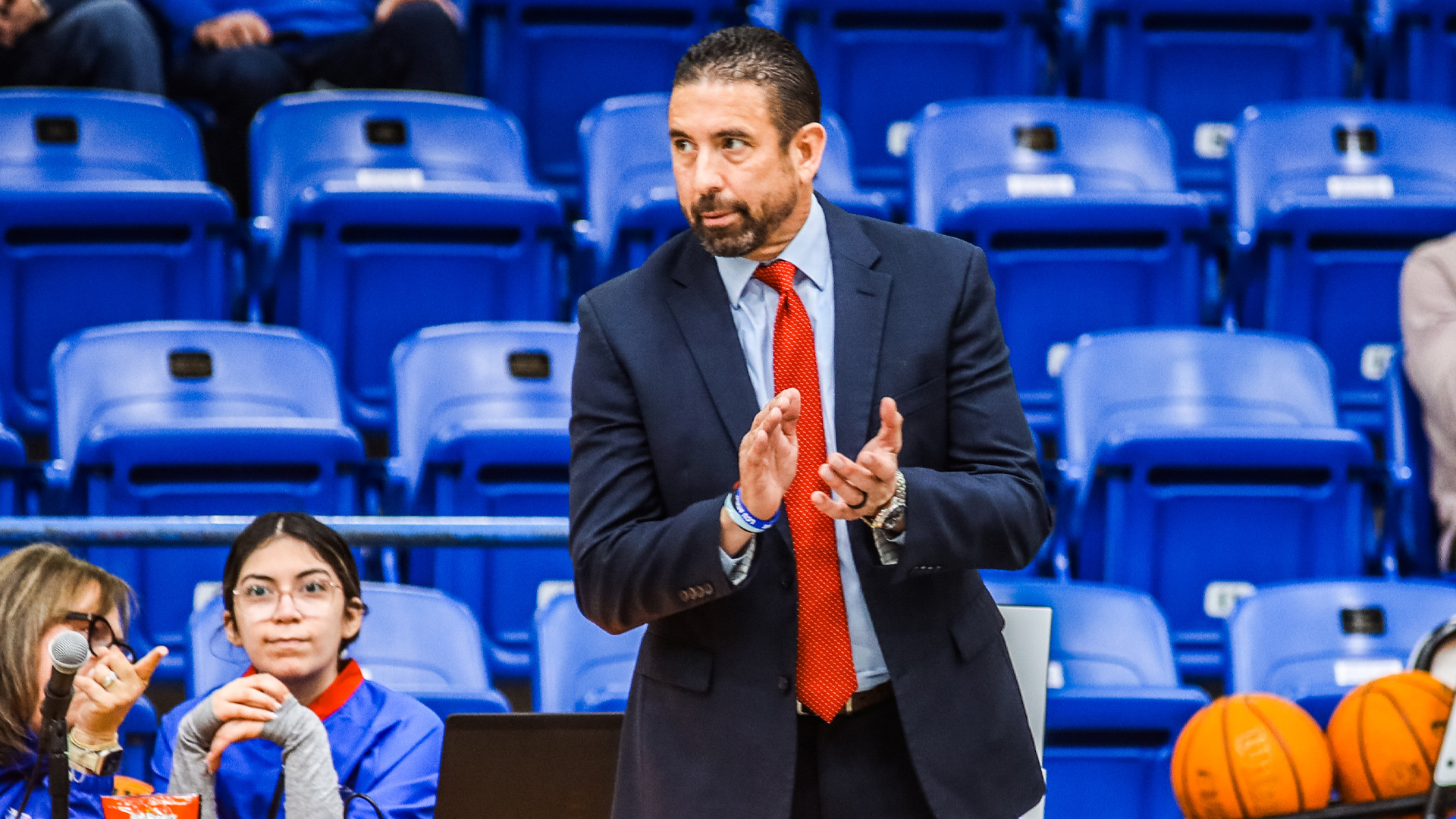LCU women's basketball head coach in dark suit with red tie clapping hands while coaching from sideline. Blue arena seating in background with spectators. Coach displays animated expression while directing team during game. Orange basketballs visible on scorer's table in lower right corner. Professional coaching moment showing engaged sideline leadership and instruction during game action