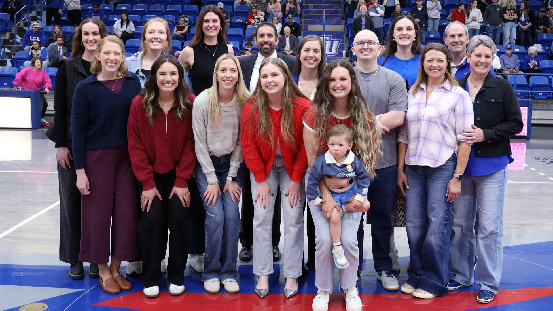 LCU women's basketball 2015-16 team reunion group photo at center court in Rip Griffin Center. Fifteen former players, coaches, and family members including small child posed in two rows on blue and red court. Group wearing mix of casual attire including red sweaters, black outfits, jeans, and button-up shirts. Blue arena seating filled with spectators in background. Reunion celebration during halftime showing former team members reconnecting years after playing together