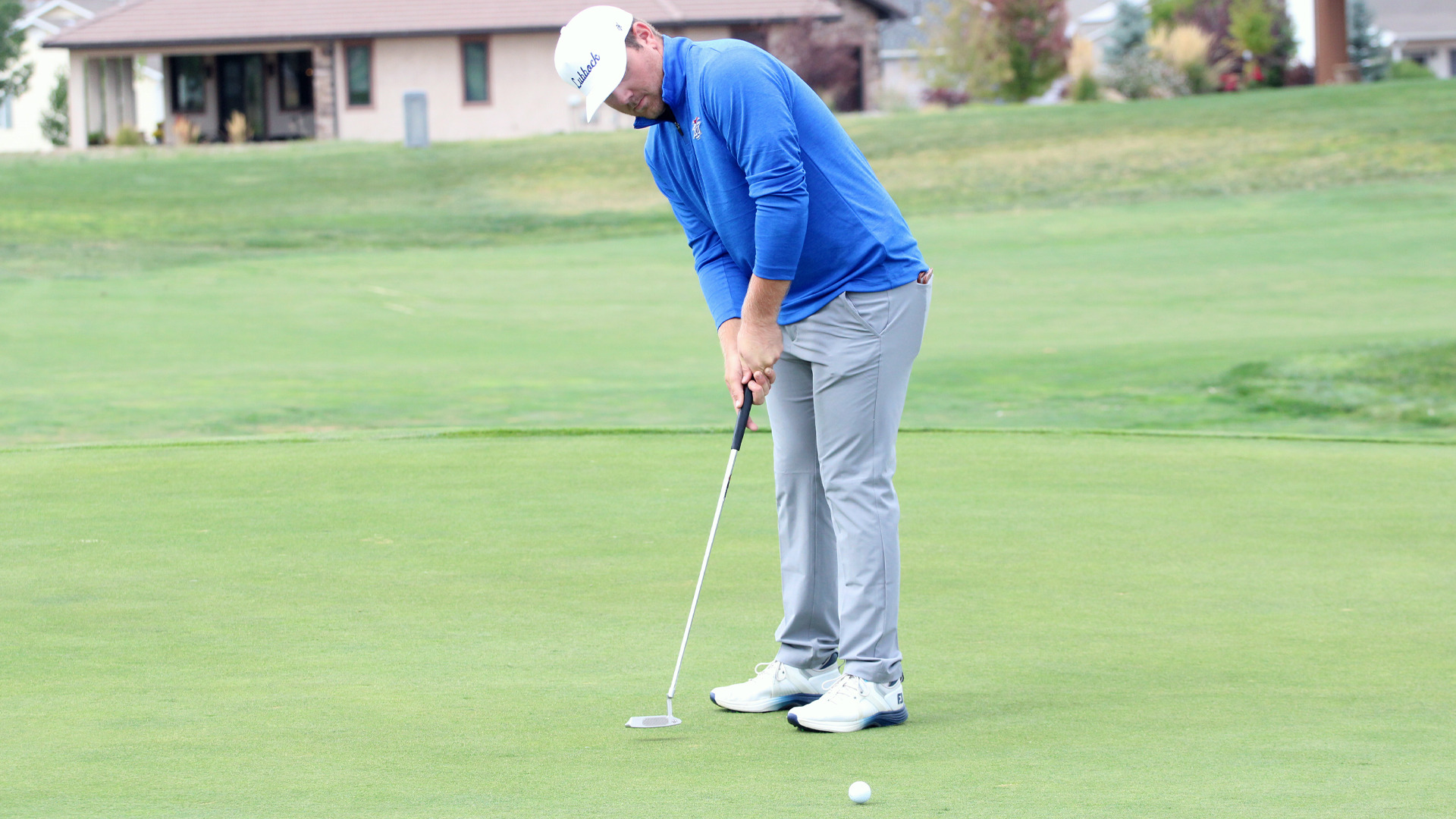 A golfer wearing a white cap, blue long-sleeve shirt, and gray pants concentrates on a putt on a smooth green. He stands in proper putting stance, leaning over his putter as a white golf ball sits just feet away on the pristine putting surface. A residential building with a pink roof is visible in the background beyond the manicured fairway