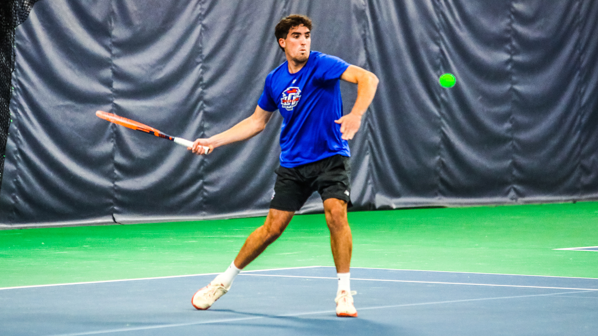 Tennis player in a blue shirt and black shorts in a defensive stance is midswing for a return volley in an indoor tennis facility