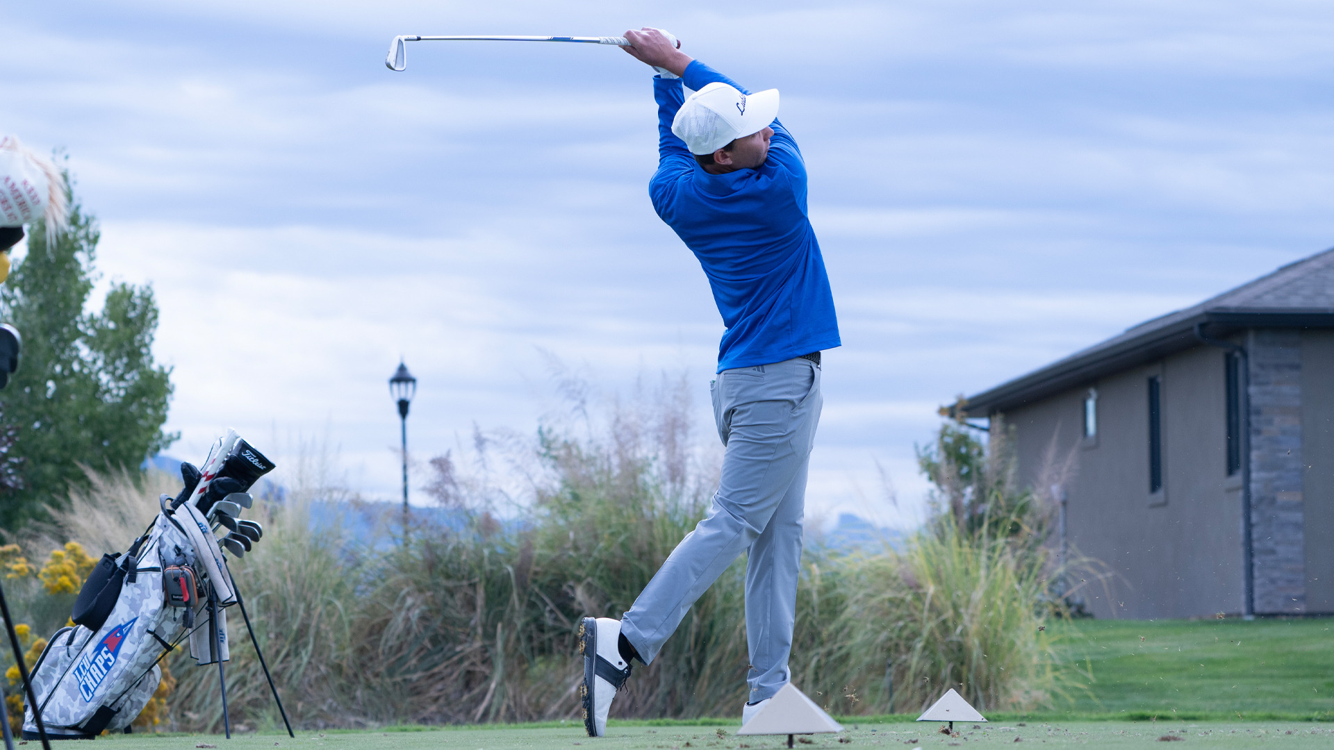 A golfer wearing a white cap, blue long-sleeve shirt, and gray pants completes his follow-through after driving off the tee box. His club is raised high overhead as he watches the ball's flight against an overcast sky. His golf bag with clubs stands nearby on the elevated tee, with natural grasses, a lamppost, and a building visible in the background of the course