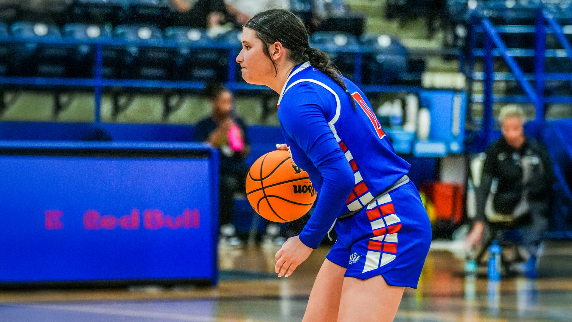 A women's basketball player in a blue jersey with red and white geometric patterns holds an orange Wilson basketball while in a ready stance on the court. She focuses intently on the play with her hair pulled back in a ponytail. The arena features blue seating and railings in the background, with spectators and coaches visible along the sidelines during game action.