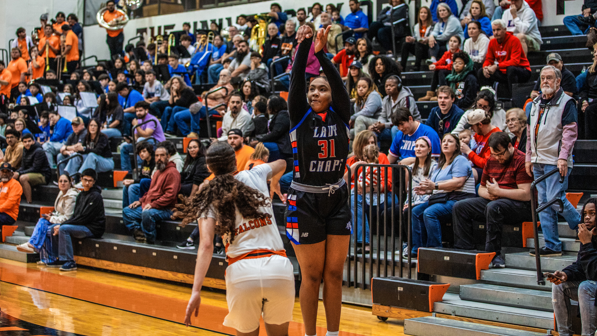 A women's basketball player wearing number 31 in a black Lady Chaps jersey with blue and red trim shoots a jump shot in front of a packed crowd. An opponent in a white Falcons uniform defends nearby on the wooden court. Spectators in colorful clothing, including many in orange, blue, and red, fill the bleachers behind the action during this away game atmosphere.