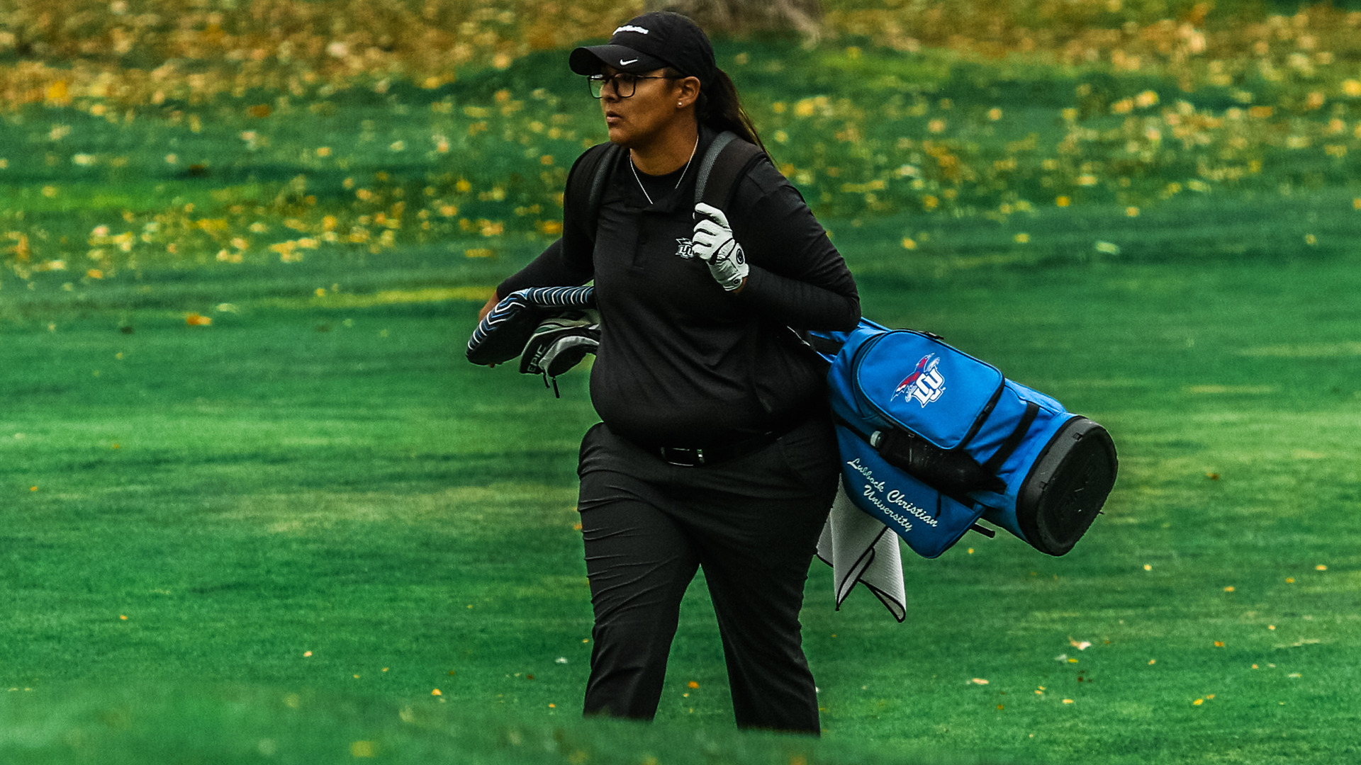 A Lubbock Christian University women's golfer walks the fairway in full black athletic attire and a golf glove, carrying clubs while slinging a blue LCU golf bag over her shoulder. Fall foliage is visible in the background on the green course.