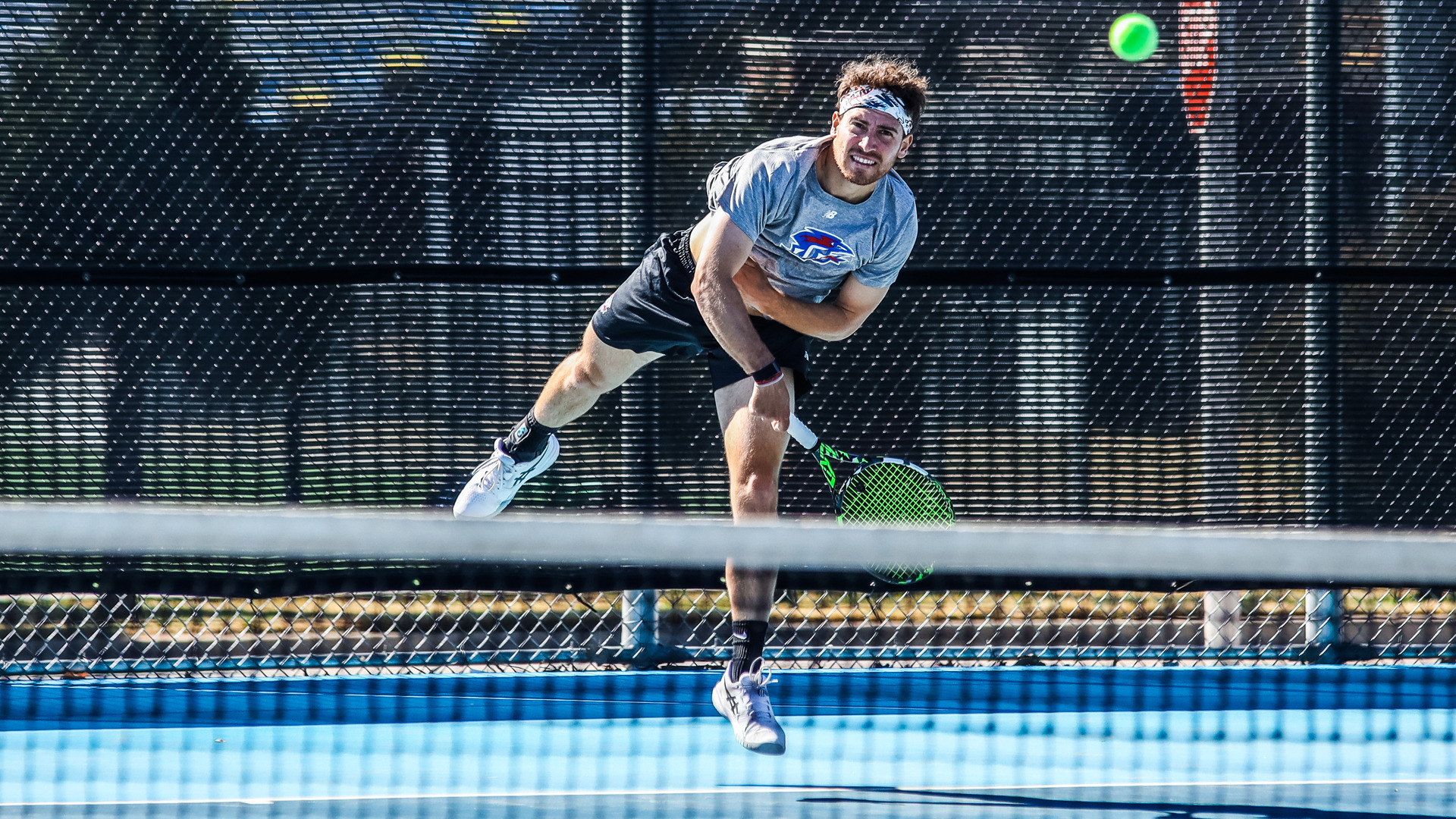 An LCU men's tennis player in a grey New Balance shirt and headband leaps into a powerful serve over the net on an outdoor blue court