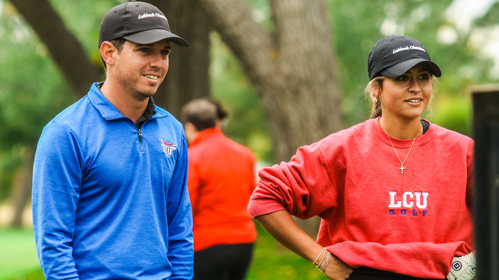Two LCU golf team members standing on course wearing black 'Lubbock Christian' caps. Left person wears blue quarter-zip pullover with LCU logo. Right person wears red 'LCU GOLF' sweatshirt with cross necklace and bracelets. Both smiling, watching play with blurred golfer in orange shirt visible in background. Lush green trees and foliage create natural outdoor setting on sunny day