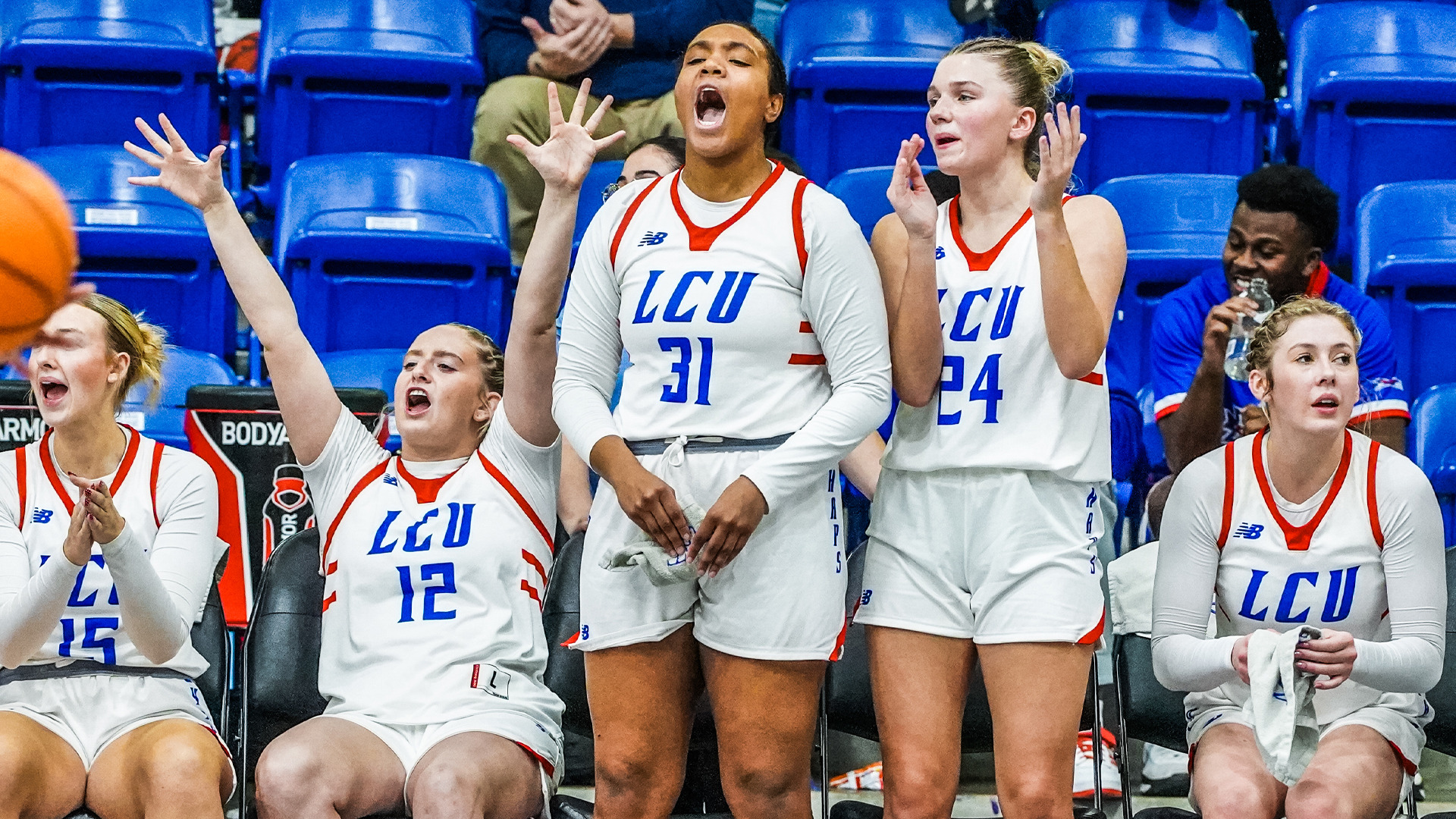 LCU women's basketball bench celebrating energetically during game. Players in white uniforms with red trim and 'LCU' in blue letters showing excited reactions. Center players number 31 and 24 standing with raised hands cheering loudly. Seated players number 12 on left with arms raised, and players on right watching intently. Blue arena seating in background. Basketball visible in motion on left edge. Team displays enthusiastic support from sideline