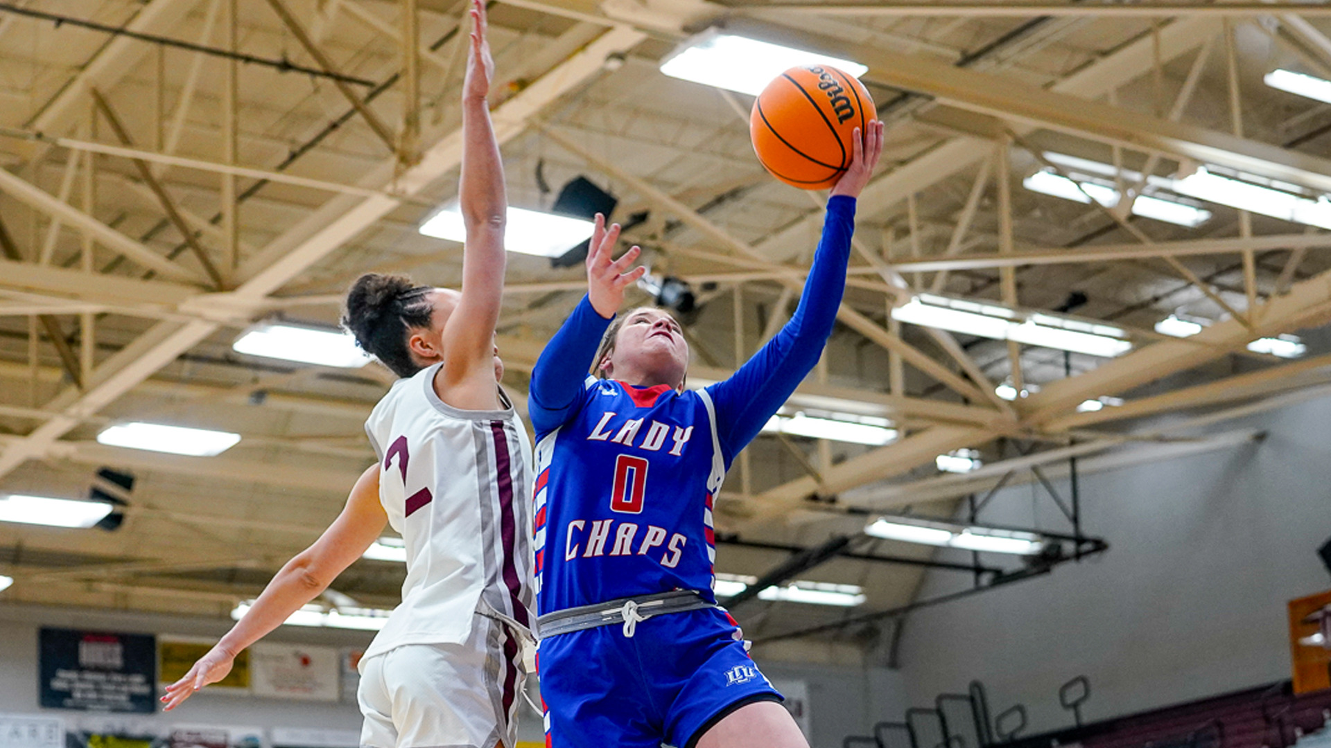 LCU women's basketball player number 0 in blue long-sleeve 'LADY CHAPS' uniform with red trim driving to basket for layup with Wilson basketball, extending right arm upward. Defended by opponent number 2 in white and maroon uniform with arm raised. Indoor arena with tan exposed ceiling beams and fluorescent lighting visible in background. Dynamic game action showing offensive move toward basket