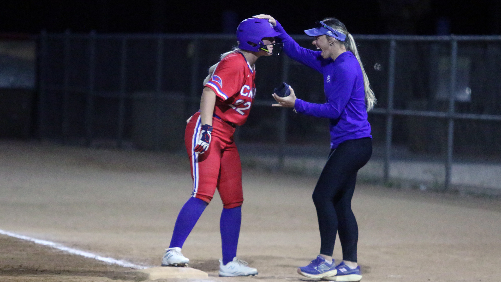 LCU softball night game showing coach in purple long-sleeve top, black pants, and blue visor celebrating with player number 22 in red 'CHAPS' uniform and blue batting helmet at base. Coach reaches toward player's helmet in congratulatory gesture. Dark night sky background with chain-link fence and field lights visible. Coaching moment capturing successful play during California road trip game