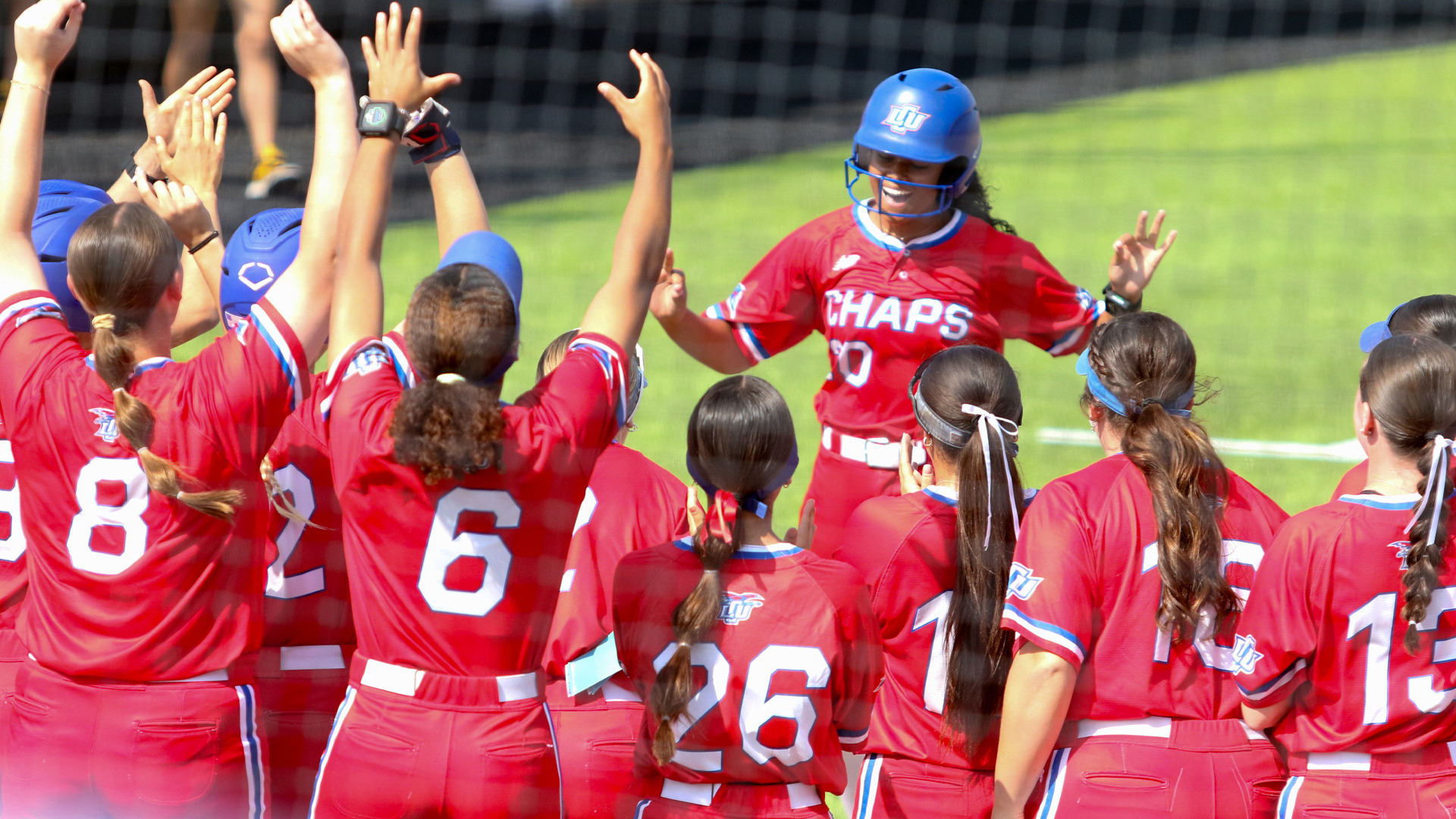 LCU Lady Chaps softball player number 20 in a red Chaps uniform and blue LCU helmet is greeted by teammates with raised hands at home plate after scoring a run against Cameron
