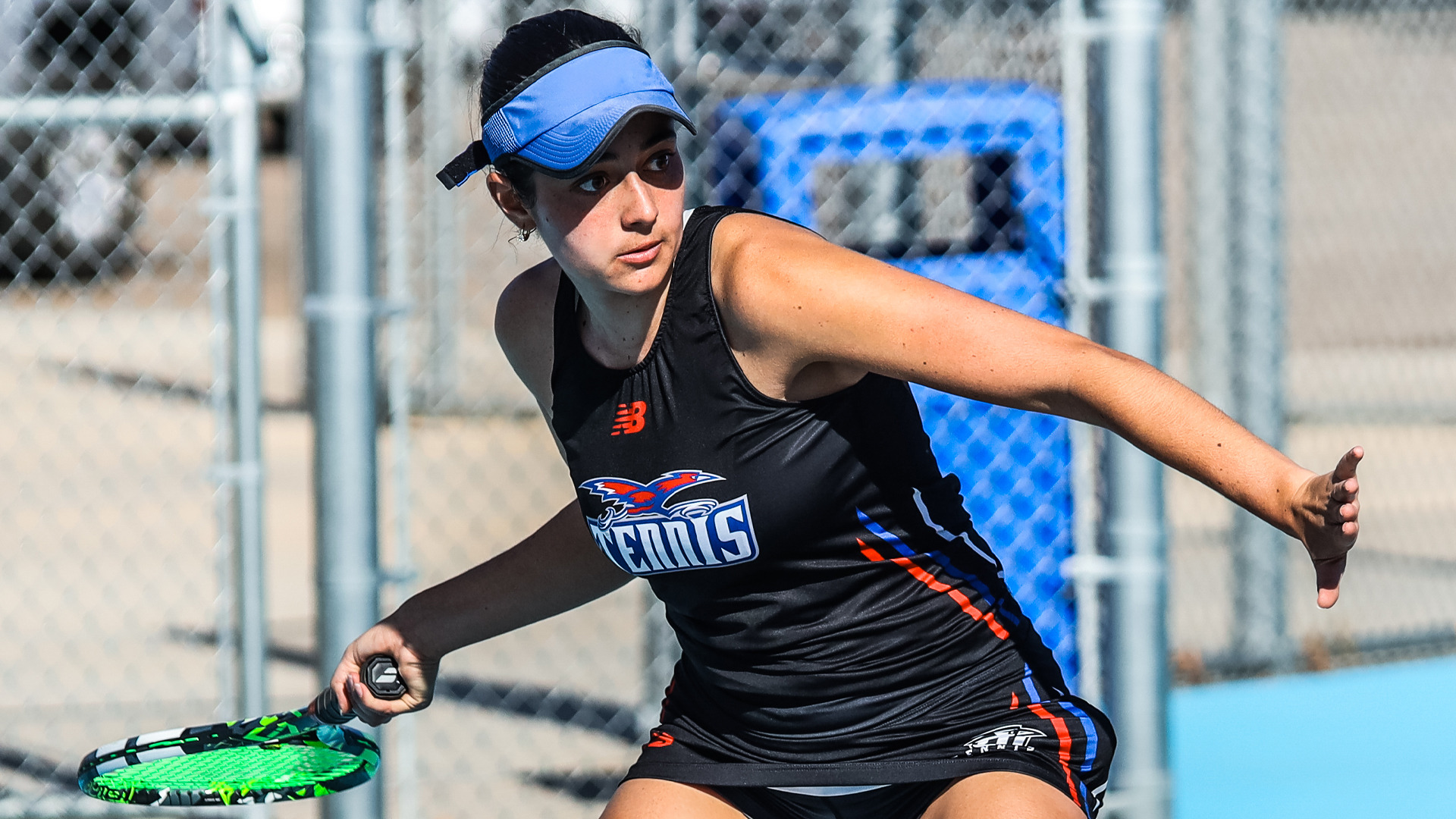 An LCU Lady Chaps tennis player in a black LCU Tennis uniform and blue visor lunges wide to hit a forehand on an outdoor blue court at Penny Gullo Tennis Center