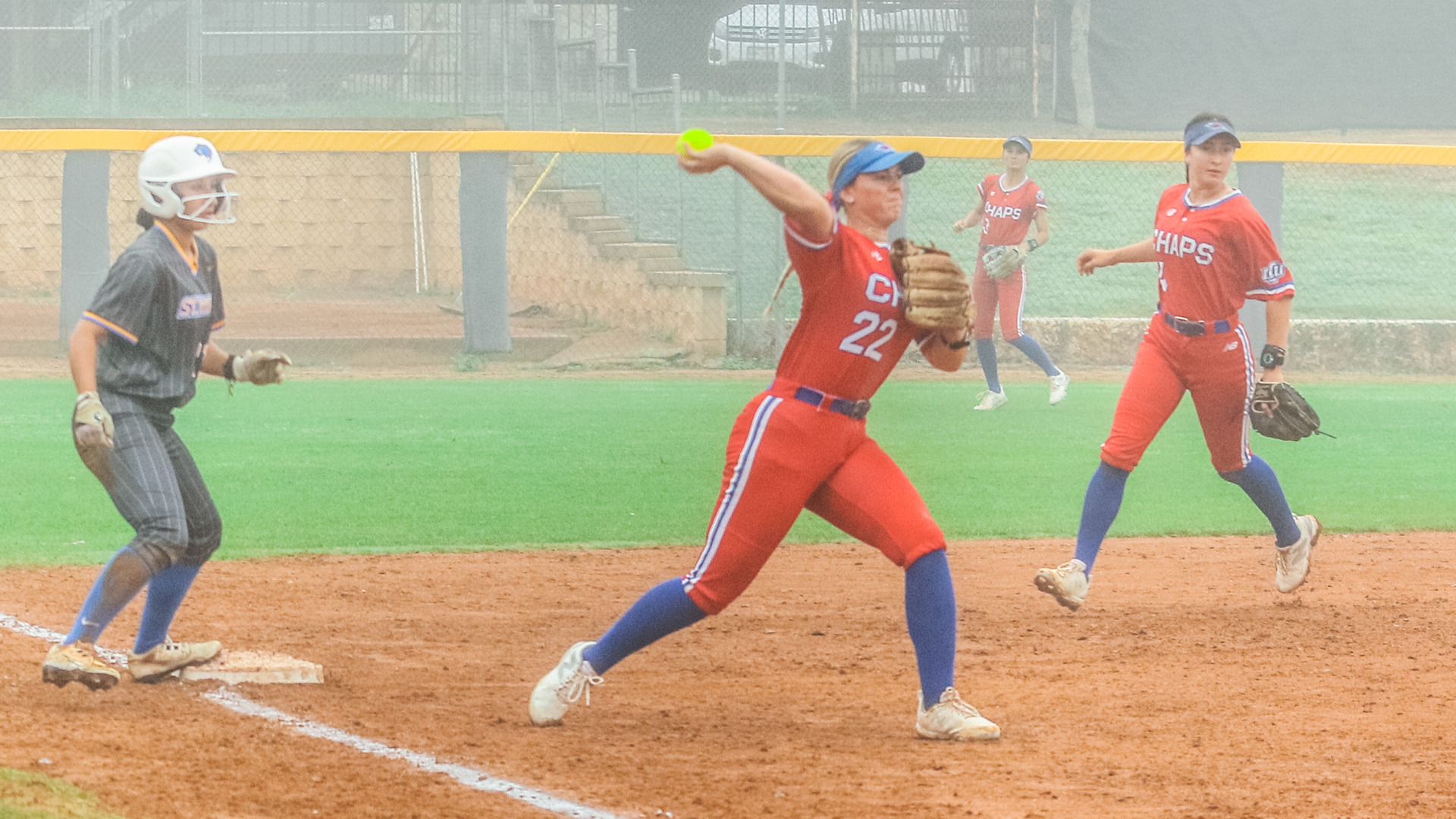 LCU softball player number 22 in a red Chaps uniform throws across the infield during a foggy game, with a baserunner and umpire visible near third base