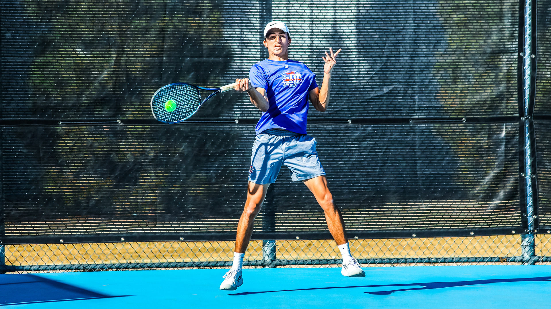 An LCU men's tennis player in a blue LCU shirt and white Nike cap hits a forehand on an outdoor blue court at Penny Gullo Tennis Center