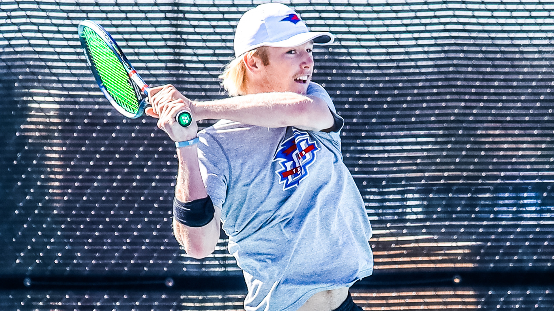 An LCU men's tennis player in a grey LCU tennis shirt and white cap follows through on a two-handed backhand on an outdoor court.