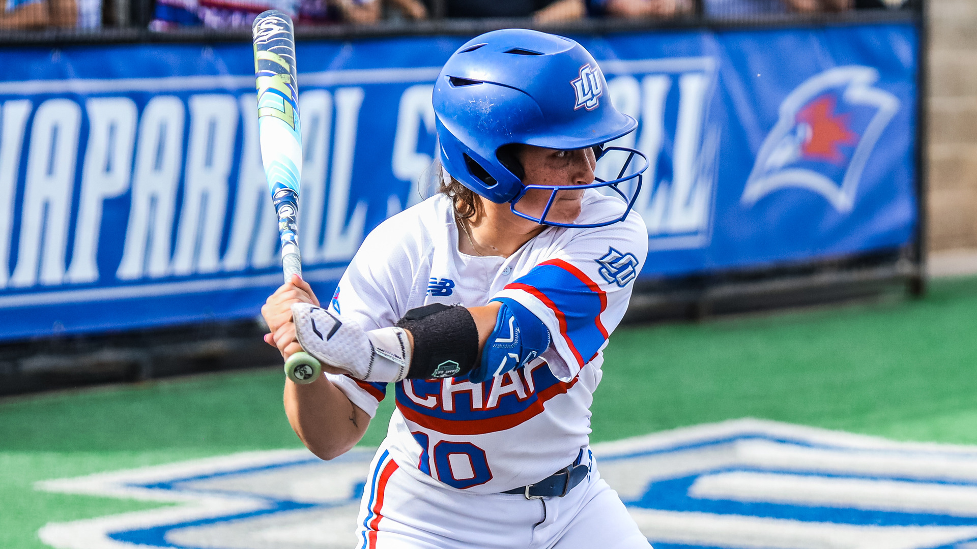 LCU softball player number 10 in a batting stance, wearing a blue helmet and white Chaps uniform with red and blue accents. She grips a colorful bat and focuses intently on the field. A blue LCU Chaparral Softball banner with the team logo is visible in the background.