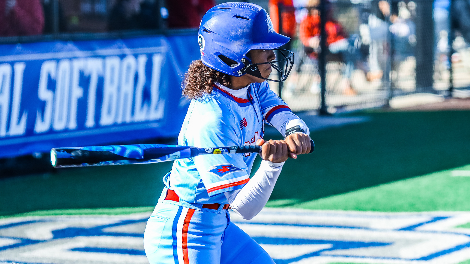 An LCU softball player in a light blue Chaps uniform with red and blue accents and blue helmet follows through on a swing. The LCU Chaparral logo is visible on her hip. A blue LCU Softball banner is displayed in the background.