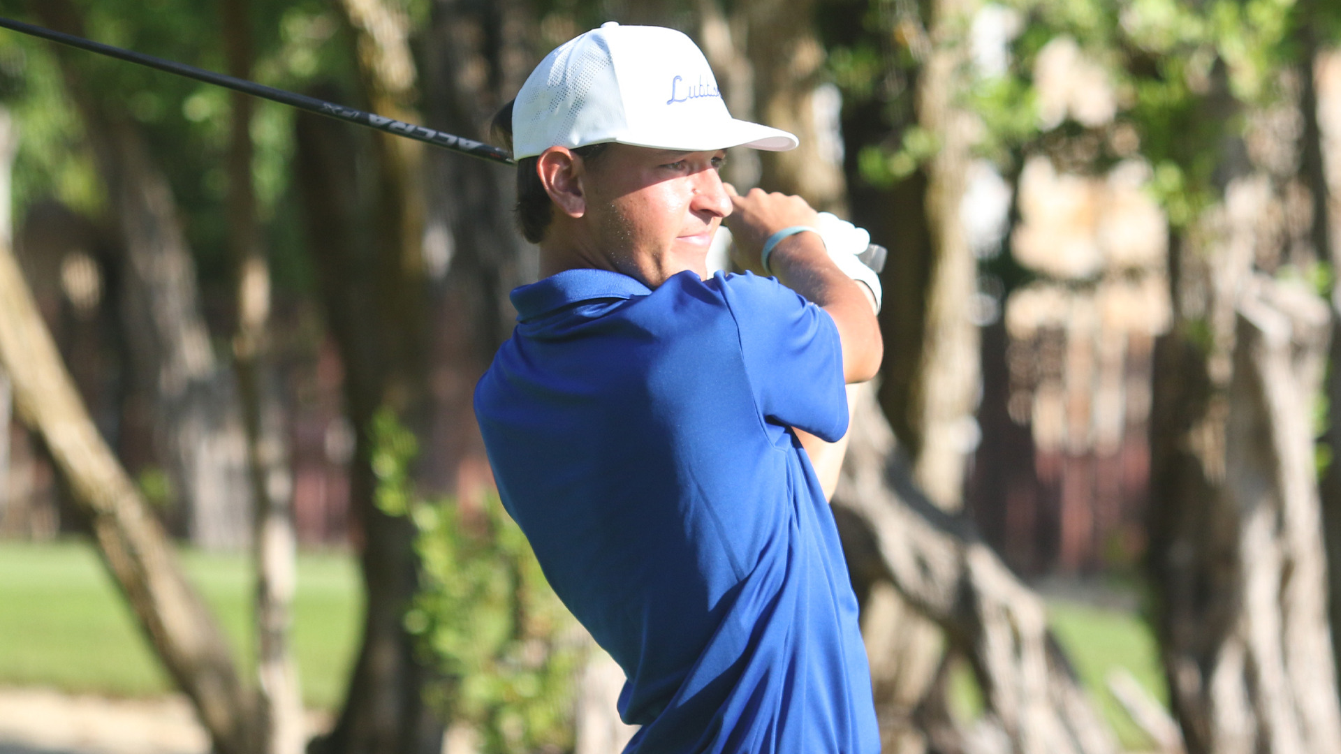 An LCU golfer in a blue polo and white LCU cap follows through on a drive, watching the ball against a tree-lined background.