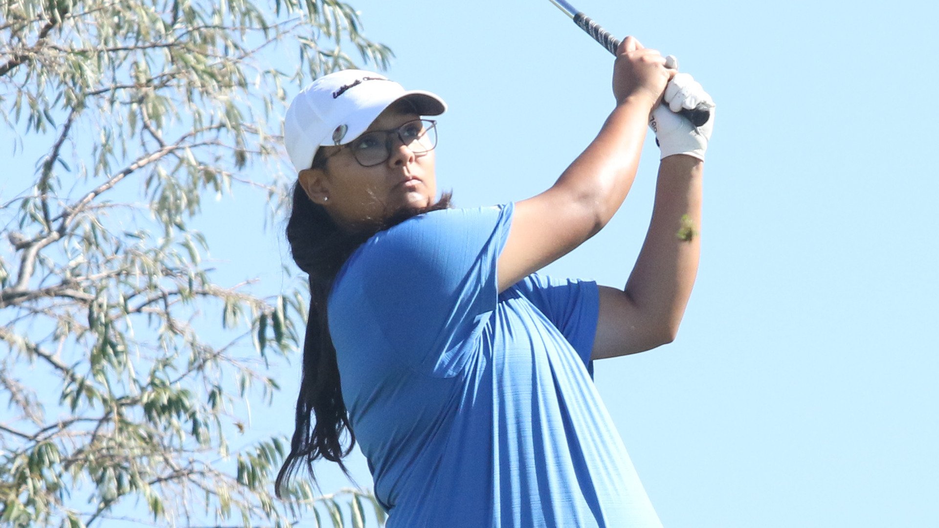 An LCU golfer in a blue polo, white cap, and glasses follows through on a drive against a clear blue sky with tree branches visible in the background.