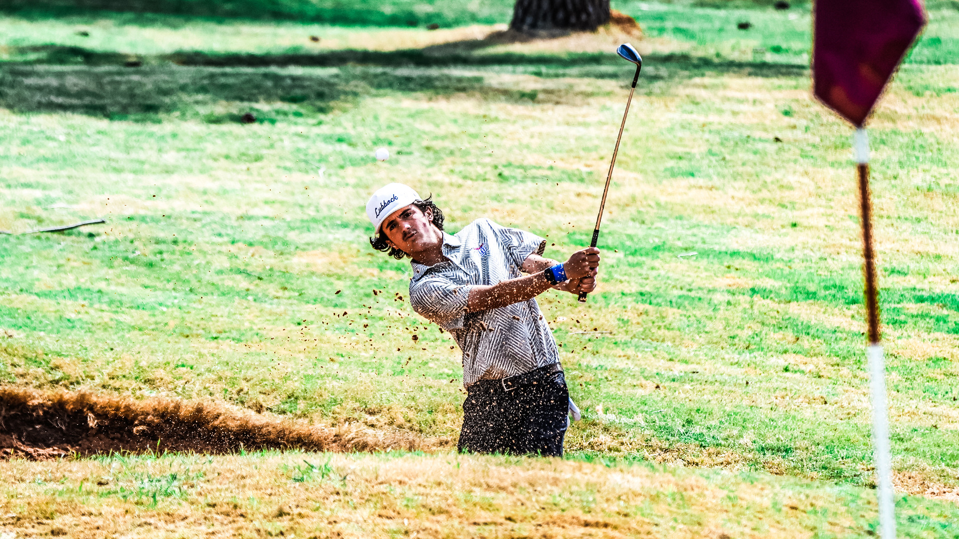 An LCU golfer in a white Lubbock cap blasts out of a bunker, sending sand and dirt flying, with a flagstick visible in the foreground.