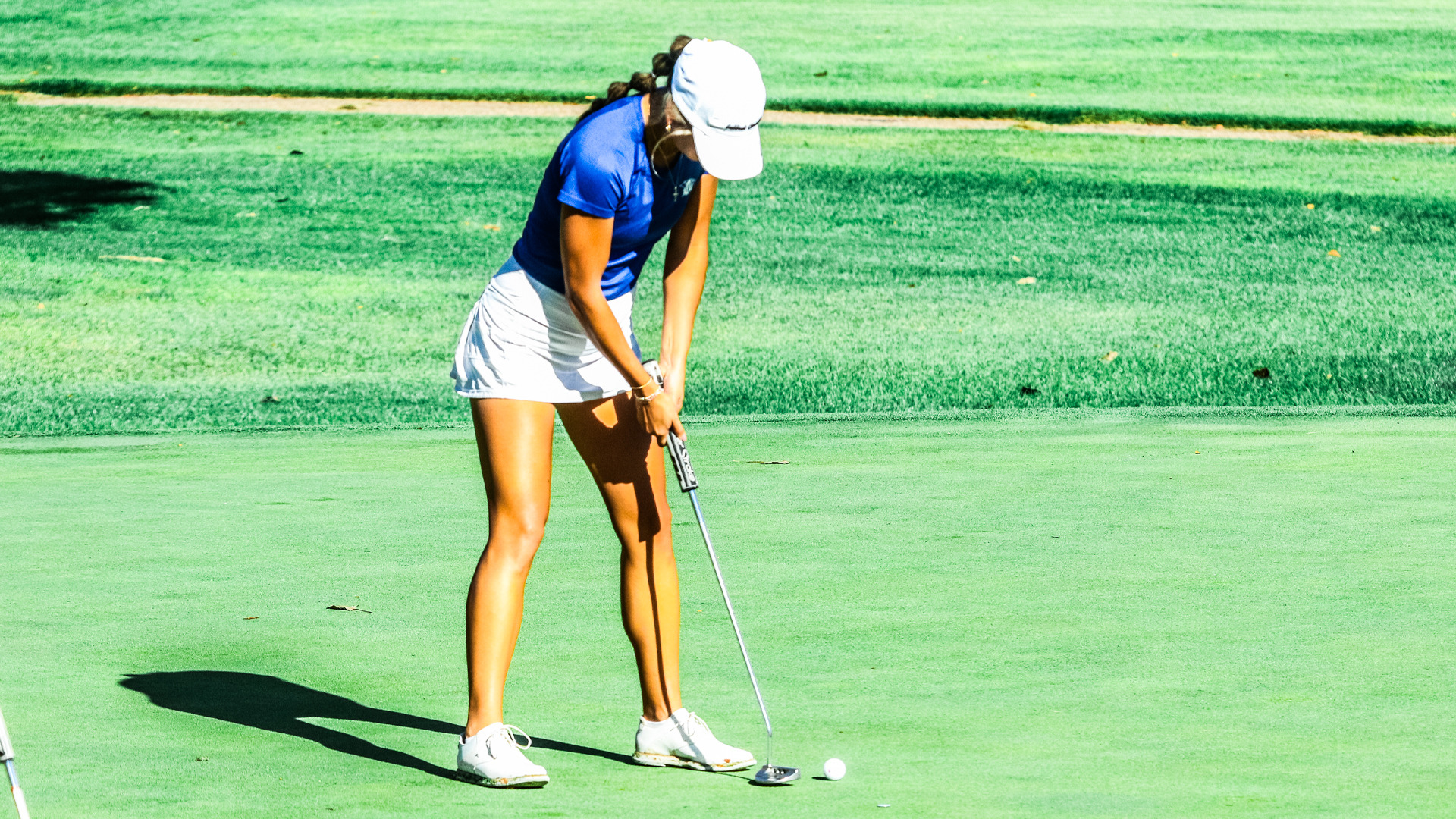 An LCU golfer in a blue polo, white skirt, and white cap lines up a putt on a sunlit green.