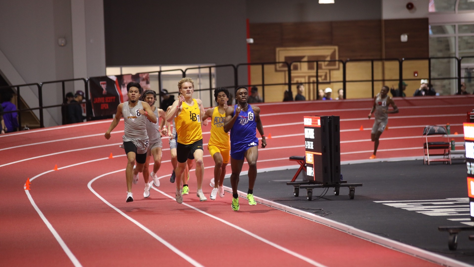 This is a photo of LCU track athlete Matthew Gordon (blue uniform) running a race on a red track at the Sport Performance Center at Texas Tech. 