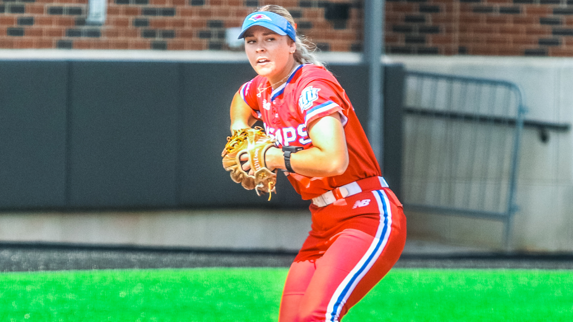 An LCU softball infielder in a red Chaps uniform and blue visor fields a ball with her glove in a ready throwing stance, with a brick wall and green turf visible in the background.