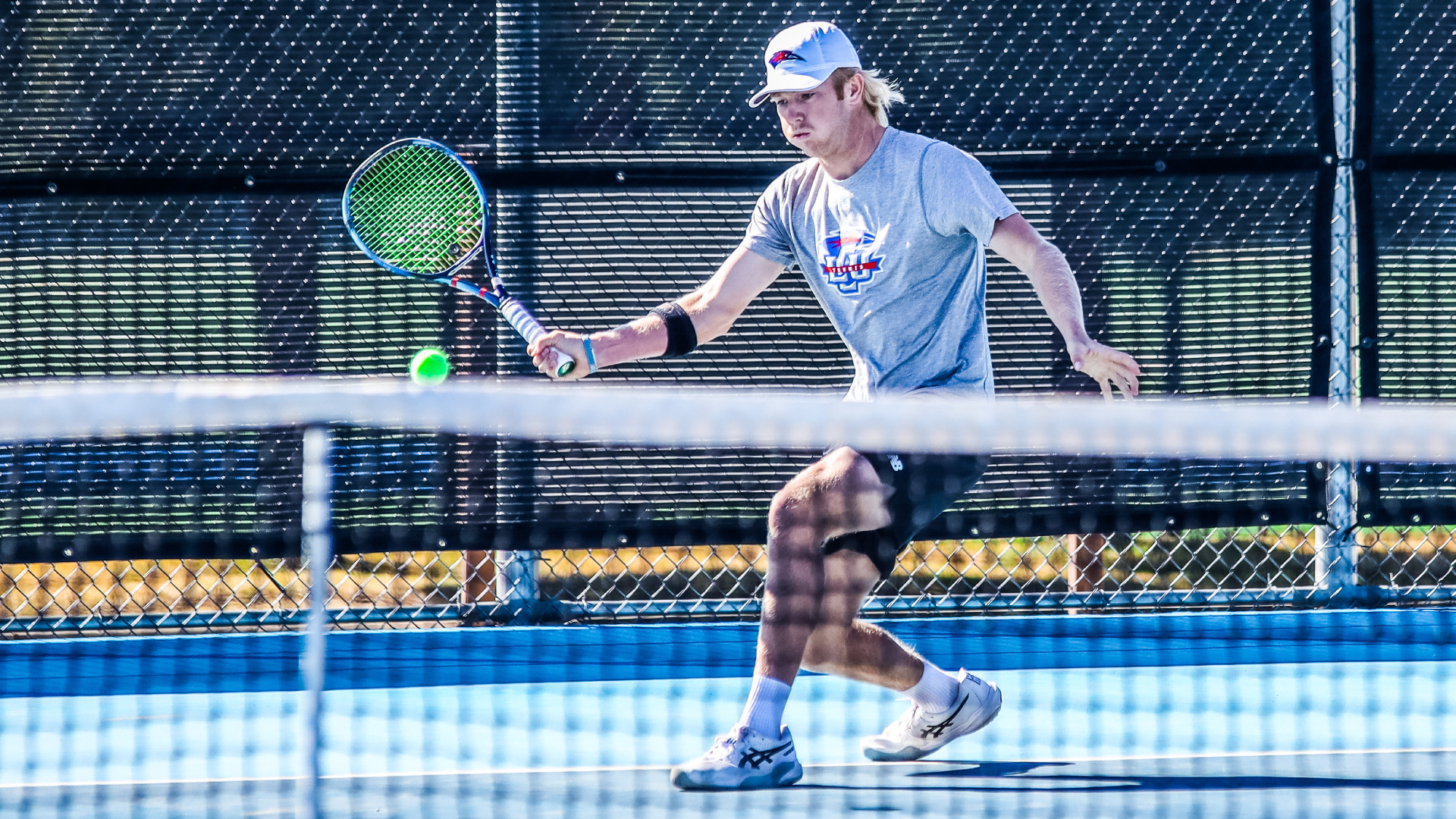 An LCU men's tennis player in a gray LCU Tennis shirt and white LCU cap lunges to strike a backhand near the net, with a tennis ball visible mid-flight against a dark windscreen background.