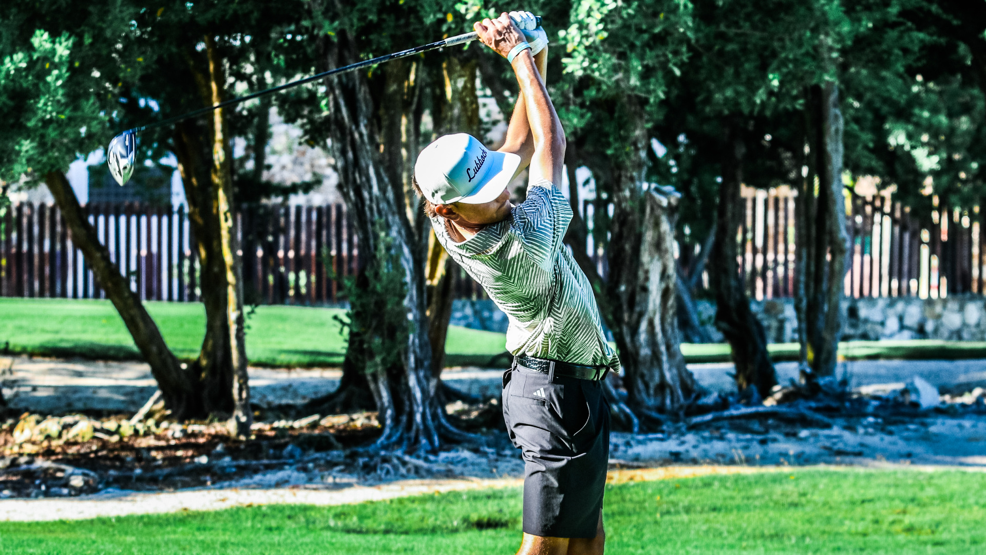 An LCU golfer in a patterned green shirt and white Lubbock cap completes a full driver swing follow-through against a tree-lined background.