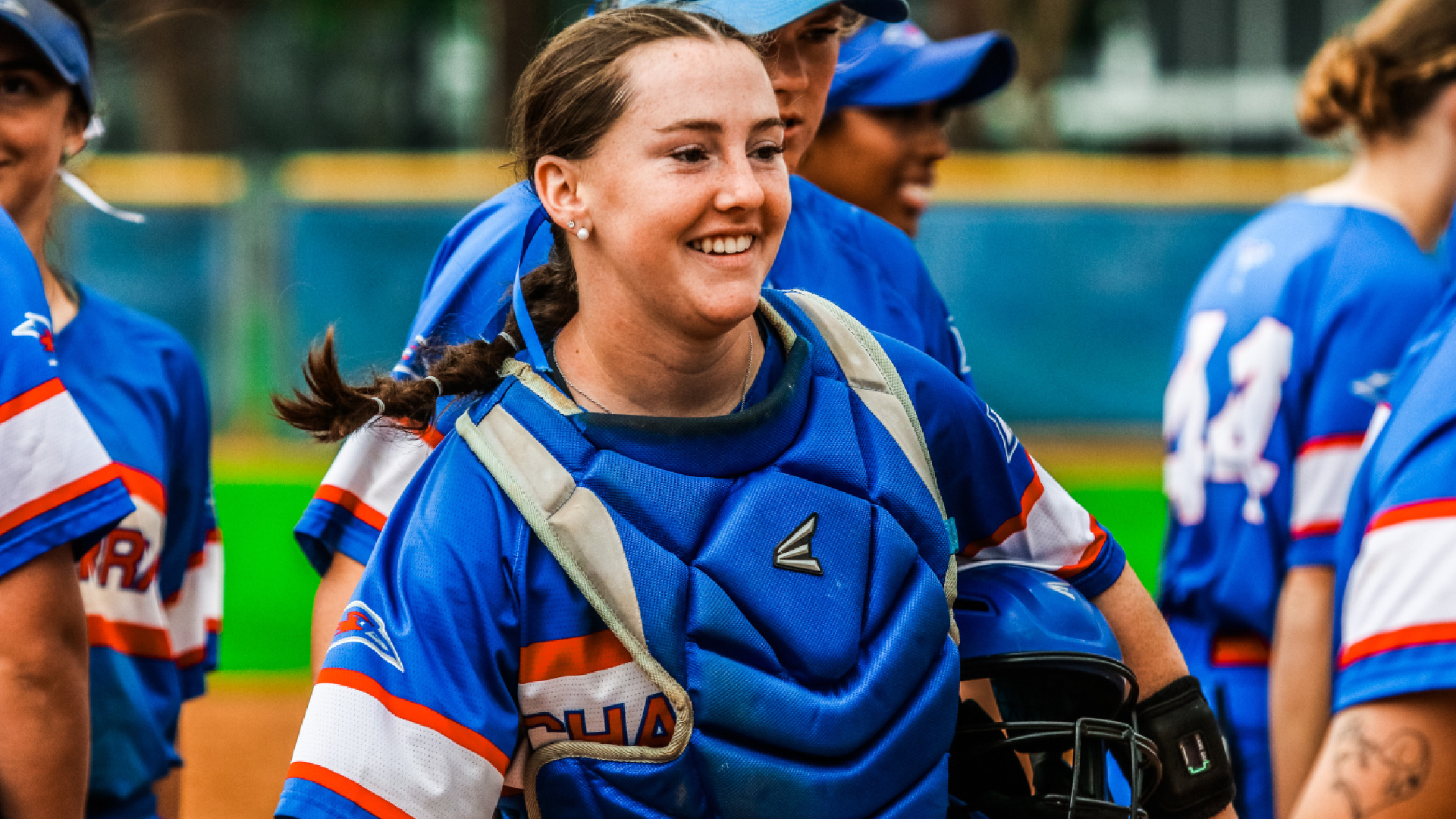 A smiling LCU softball catcher in a blue Chaps uniform and chest protector walks with teammates after a game, with green outfield turf visible in the background.