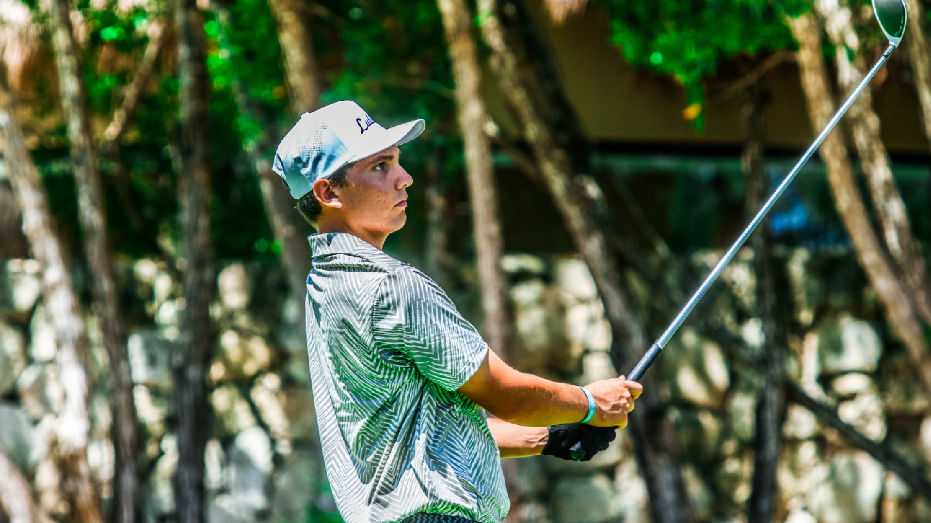 An LCU golfer in a patterned green and white shirt and white Lubbock cap watches his shot after a drive, holding the club in his follow-through against a tree-lined background.