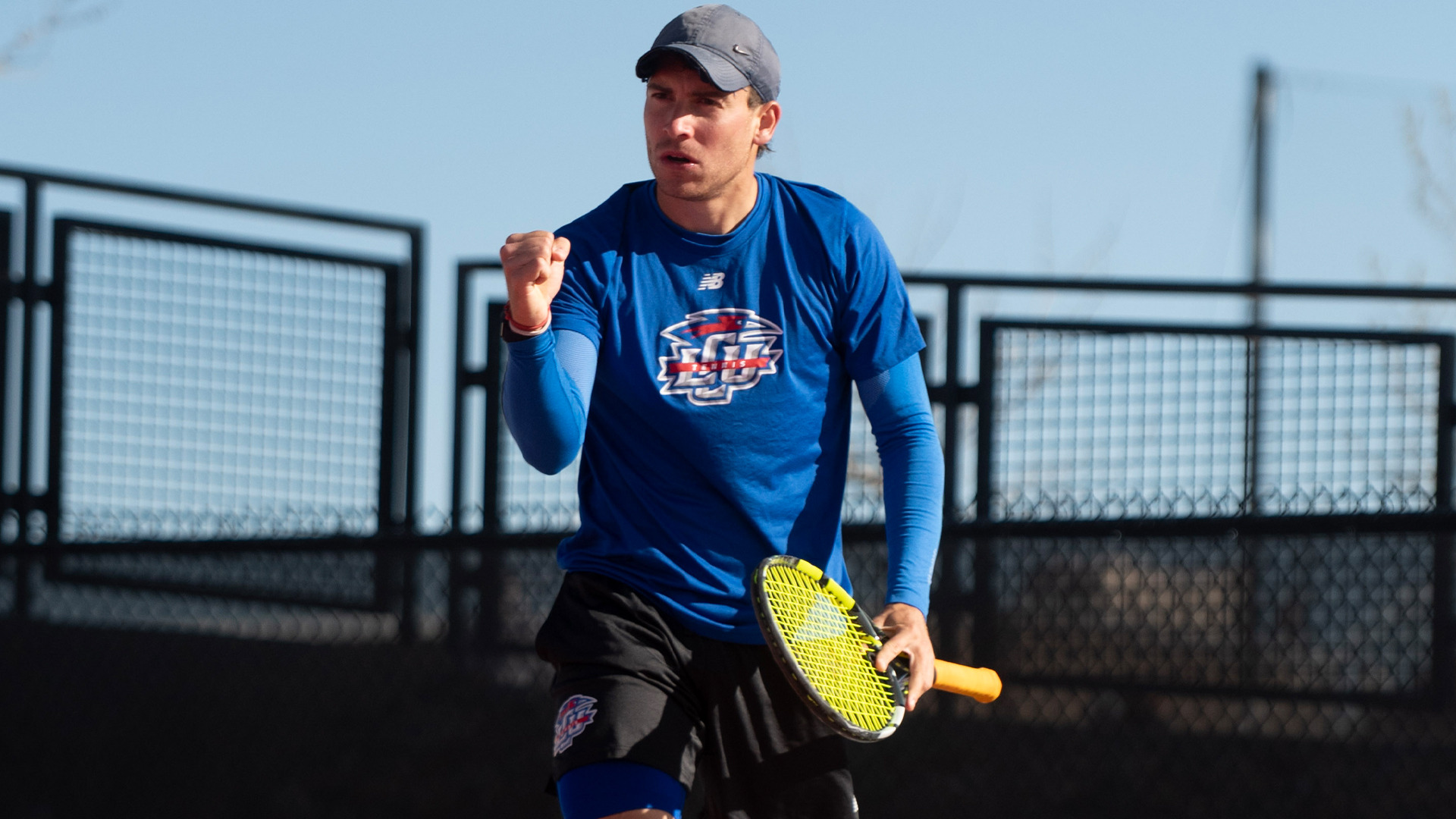 An LCU men's tennis player in a blue New Balance LCU shirt pumps his fist in celebration while holding a racket on an outdoor court