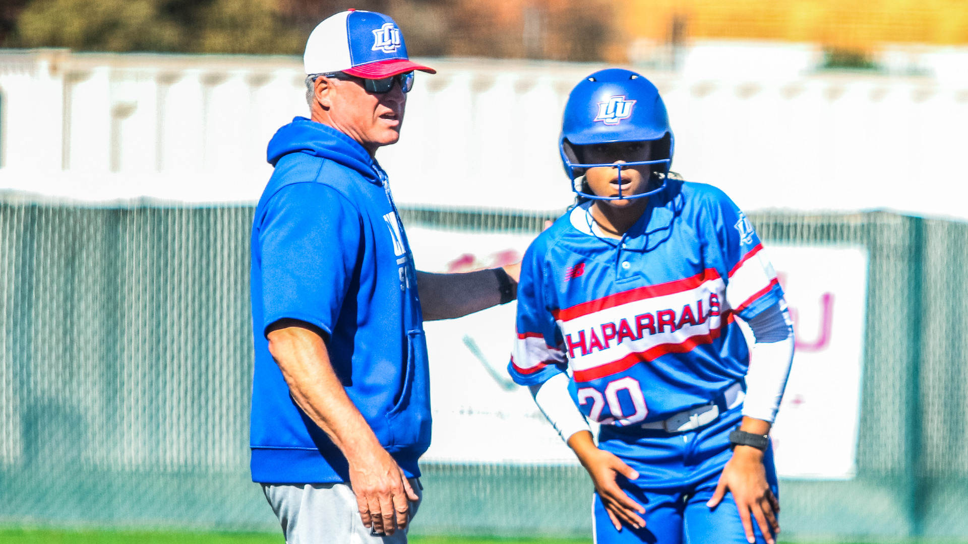 LCU softball head coach Daren Hays in a blue LCU hoodie and cap talks with player number 20 in a blue Chaparrals uniform and LCU helmet on the basepath during a game