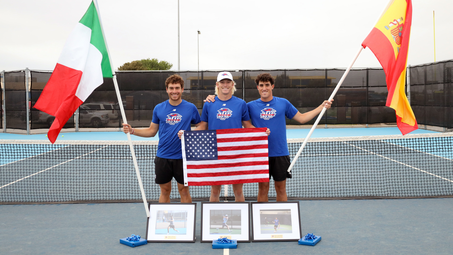 Three LCU Men's Tennis seniors pose on the tennis court during a senior day celebration. The player on the left holds the Italian flag, the center player holds an American flag, and the player on the right holds the Spanish flag. Framed photos and gift bags are displayed on the court in front of them.