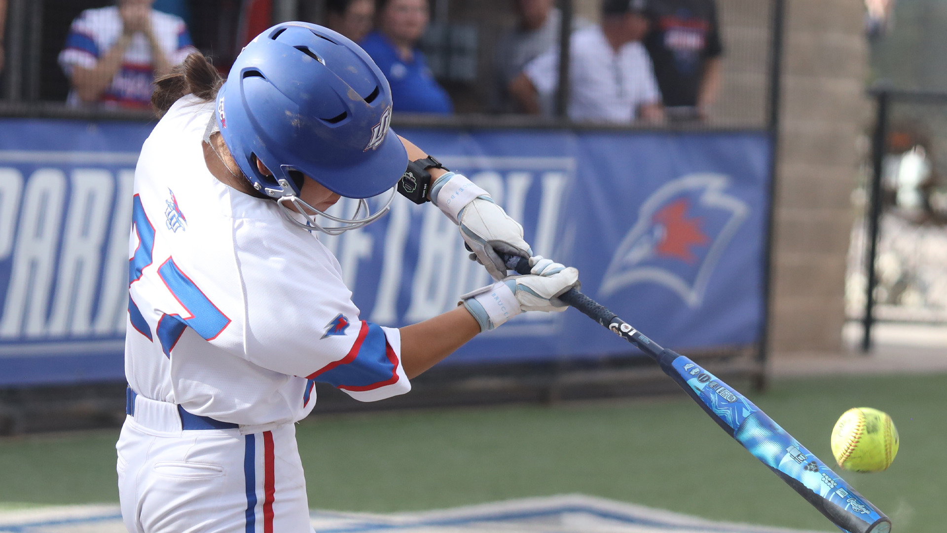 A Lady Chaps softball player in a white and blue uniform and blue helmet makes contact with a pitch, bat meeting ball at the plate. The LCU logo is visible on the helmet. Fans and a blue LCU banner are visible in the blurred background.