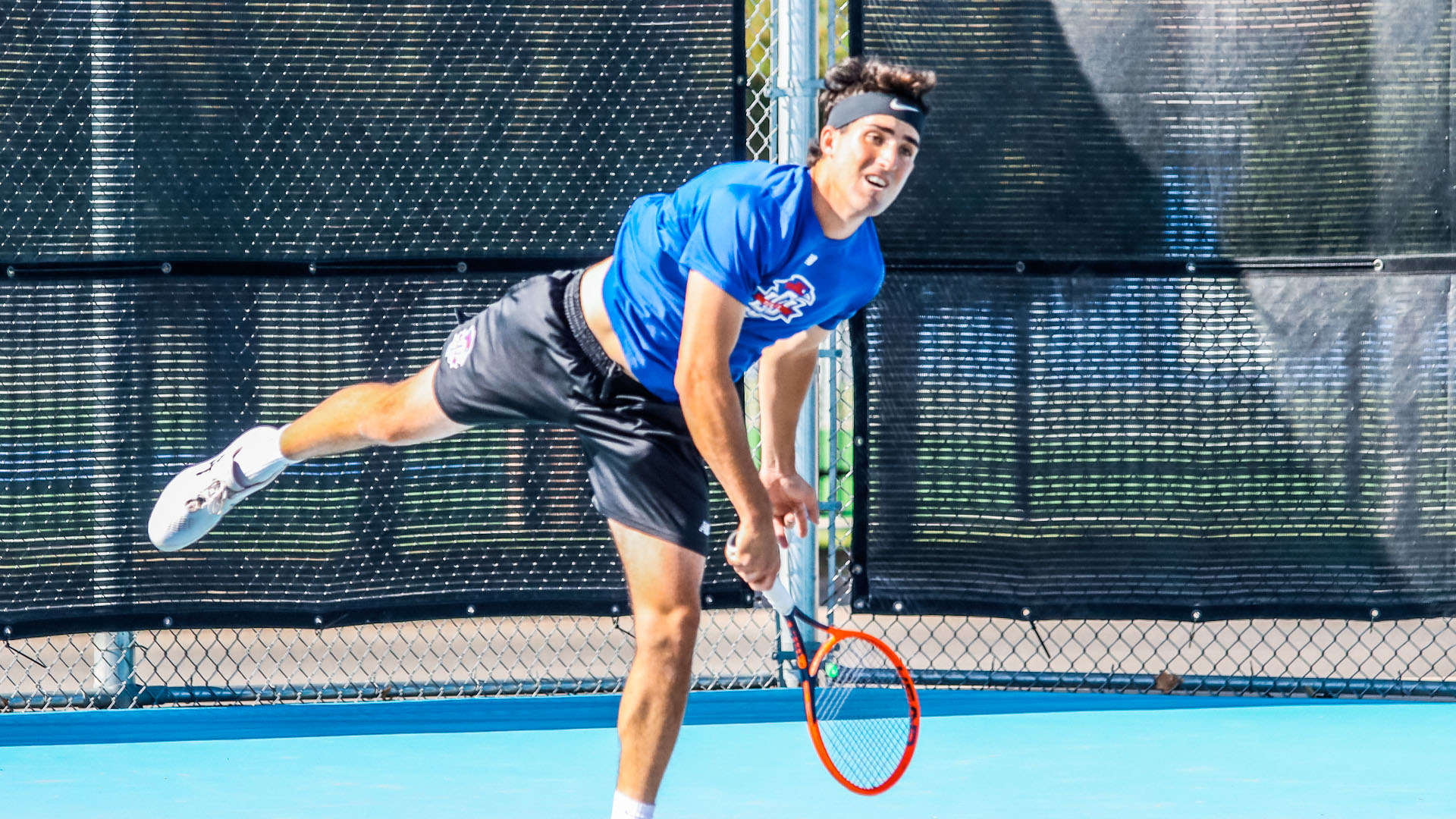 An LCU men's tennis player in a blue shirt and black headband follows through on a powerful serve on a blue outdoor court, his body leaning forward with one foot lifted off the ground.