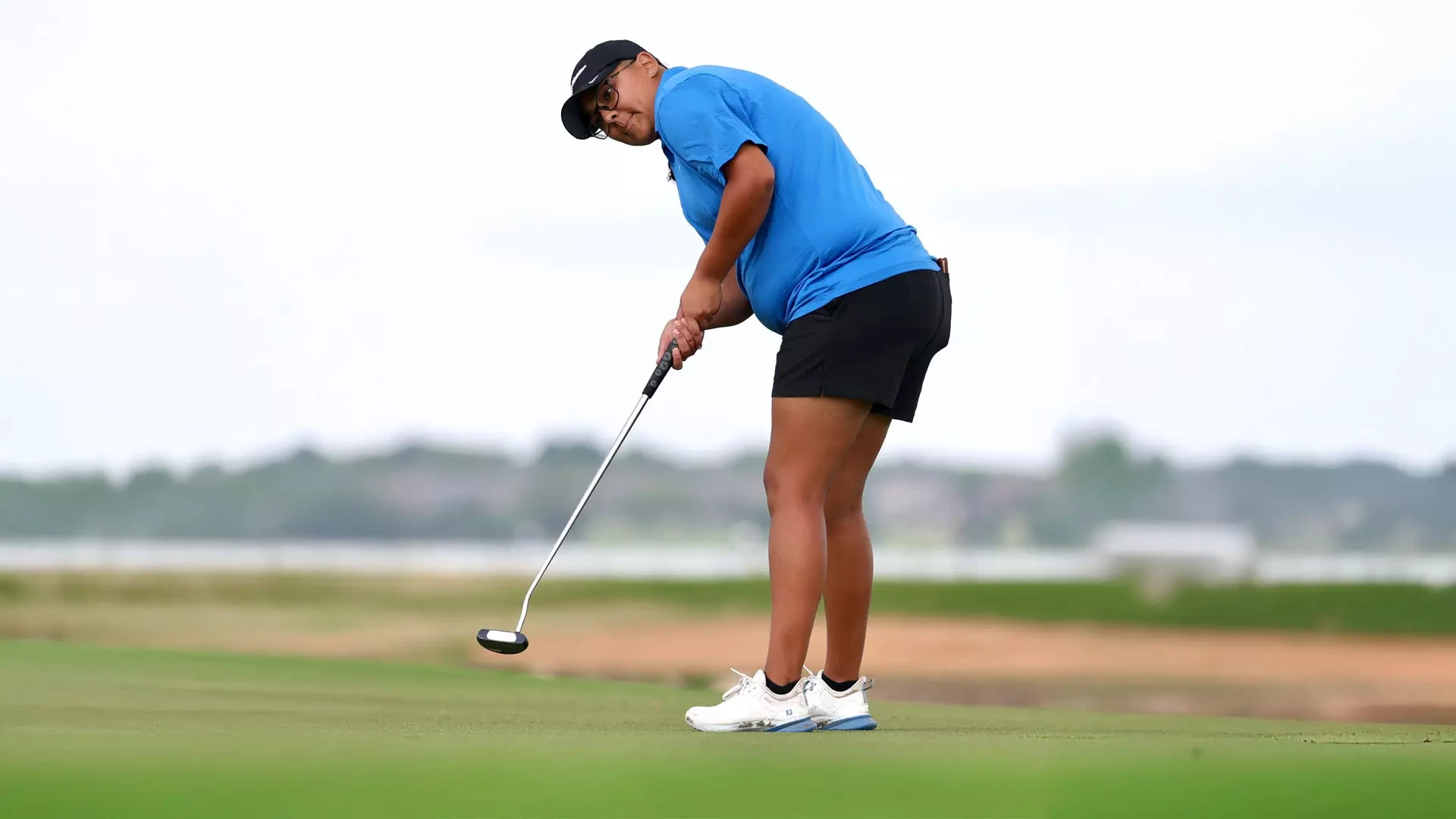 A Lady Chaps golfer in a blue shirt, black shorts, and white golf shoes leans forward to execute a putt on a green, with a blurred course and water hazard visible in the background.