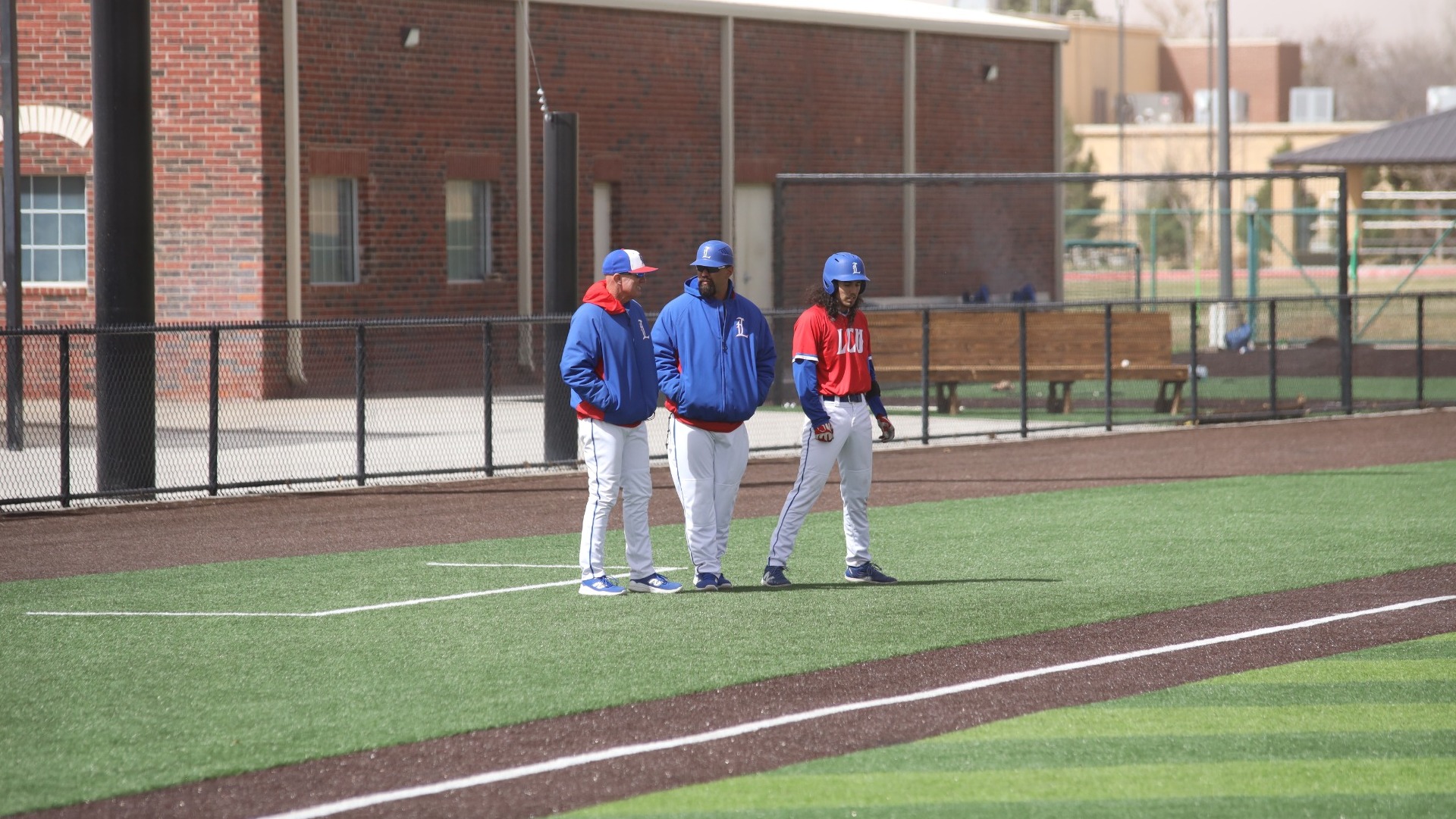 This is a photo of Lubbock Christian baseball coaches Nathan Blackwood (left, blue jacket) and Brandon Walker (middle, blue jacket) talking in the third base coaching box during a game at Hays Field. Catcher Jorge De Los Santos (red jersey, #6) stands to their right. 