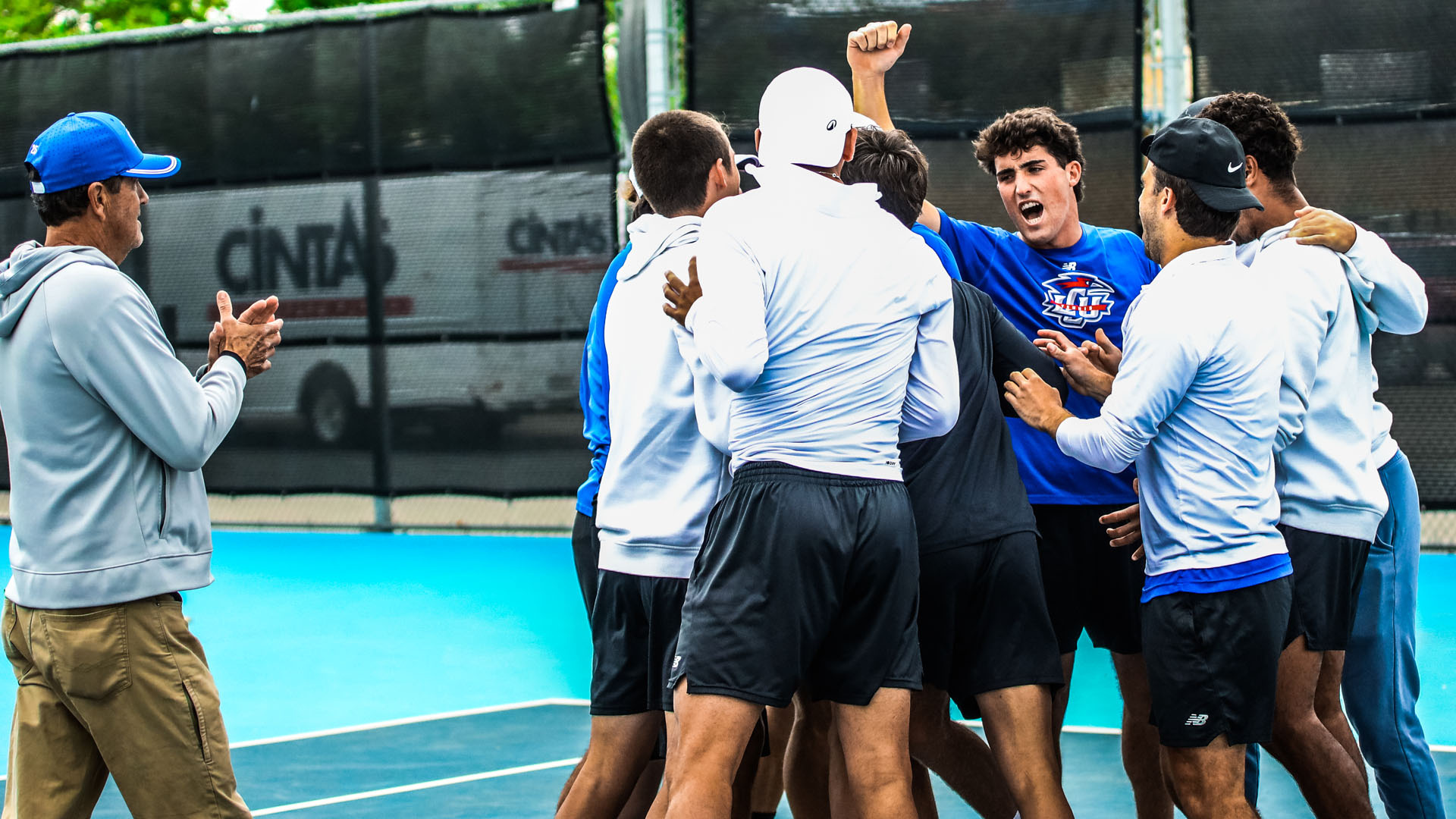 The LCU Men's Tennis team celebrates on a blue outdoor court, huddling together with one player raising his fist in the air. The coach stands to the left applauding as the team erupts in excitement.