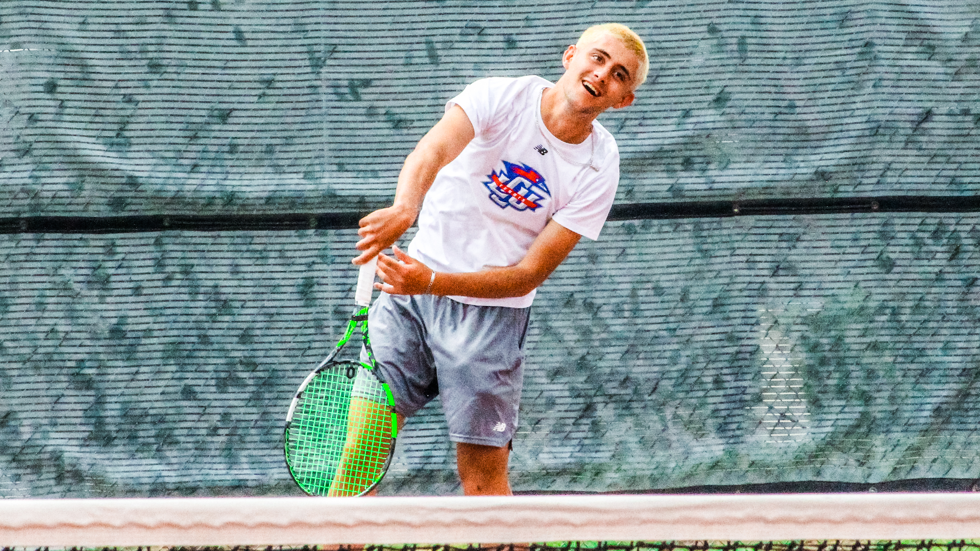An LCU men's tennis player in a white shirt and gray shorts follows through on a serve, smiling, with a dark windscreen backdrop behind him.