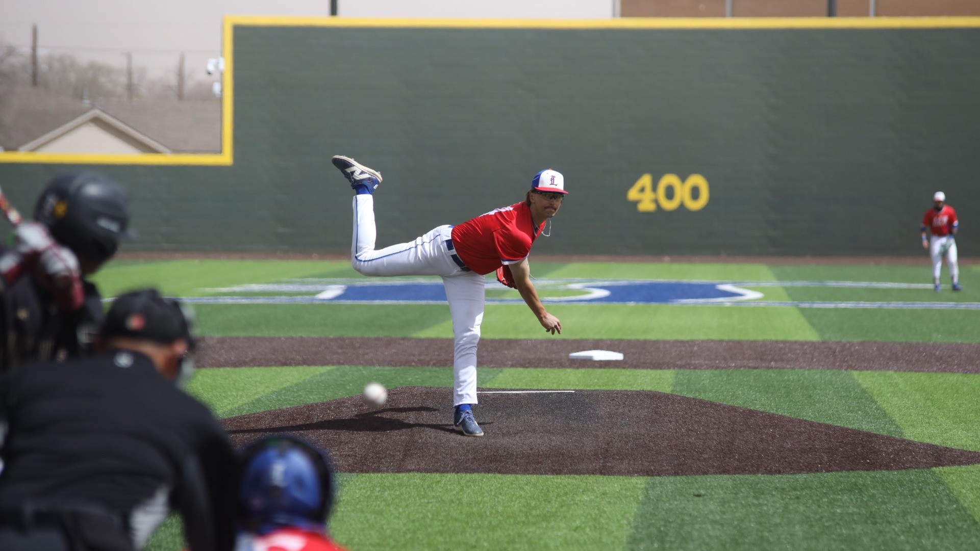 This is a photo of Lubbock Christian pitcher Bryce Nall (red jersey, #14) throwing a pitch against a Texas A&M International batter during a game at Hays Field. 
