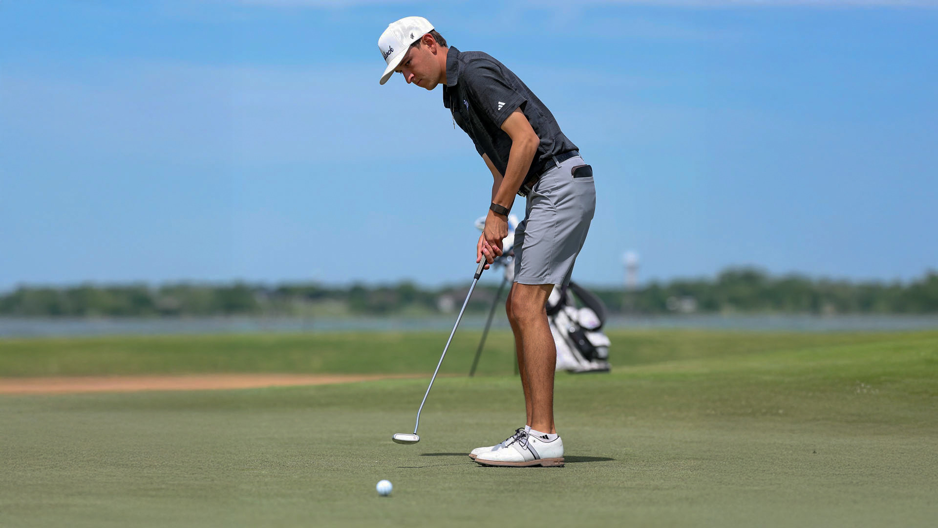 A Chaps golfer in a black polo, gray shorts, and white cap executes a putt on a sunlit green, with a golf bag, water hazard, and blue sky visible in the background.