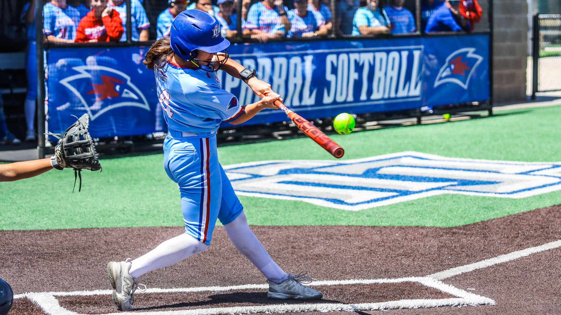 A Lady Chaps batter in a light blue uniform and blue LCU helmet makes contact with a pitch at home plate, with the LCU logo painted on the turf behind her. A catcher's mitt is visible on the left, and a crowd of fans is visible along the blue LCU Softball banner in the background.