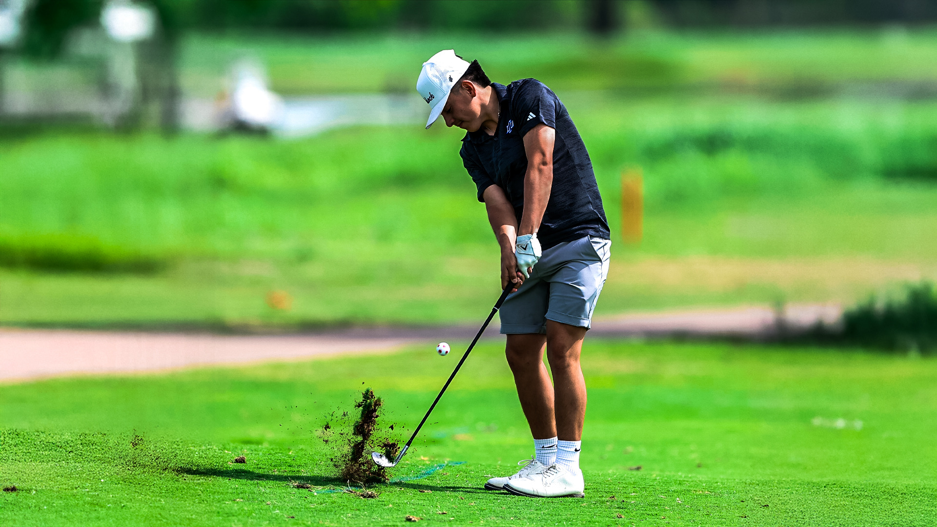 A Chaps golfer in a dark polo, gray shorts, and white cap strikes an iron shot off the fairway, sending a divot of turf into the air as the ball launches, with a lush green course background behind him.