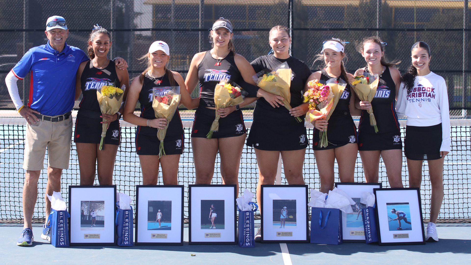 The LCU Women's Tennis seniors pose with their coach at the net during a senior day celebration. Five players in black LCU Tennis uniforms hold bouquets of flowers, with framed photos and gift bags displayed on the court in front of each of them. The coach stands on the far left, and a teammate in a Lubbock Christian Tennis sweatshirt stands on the far right.