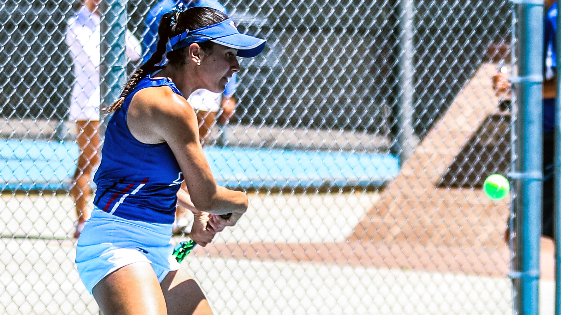 A Lady Chaps tennis player in a blue LCU uniform and blue visor strikes a backhand shot on an outdoor court, with the tennis ball visible to the right and spectators blurred in the background.