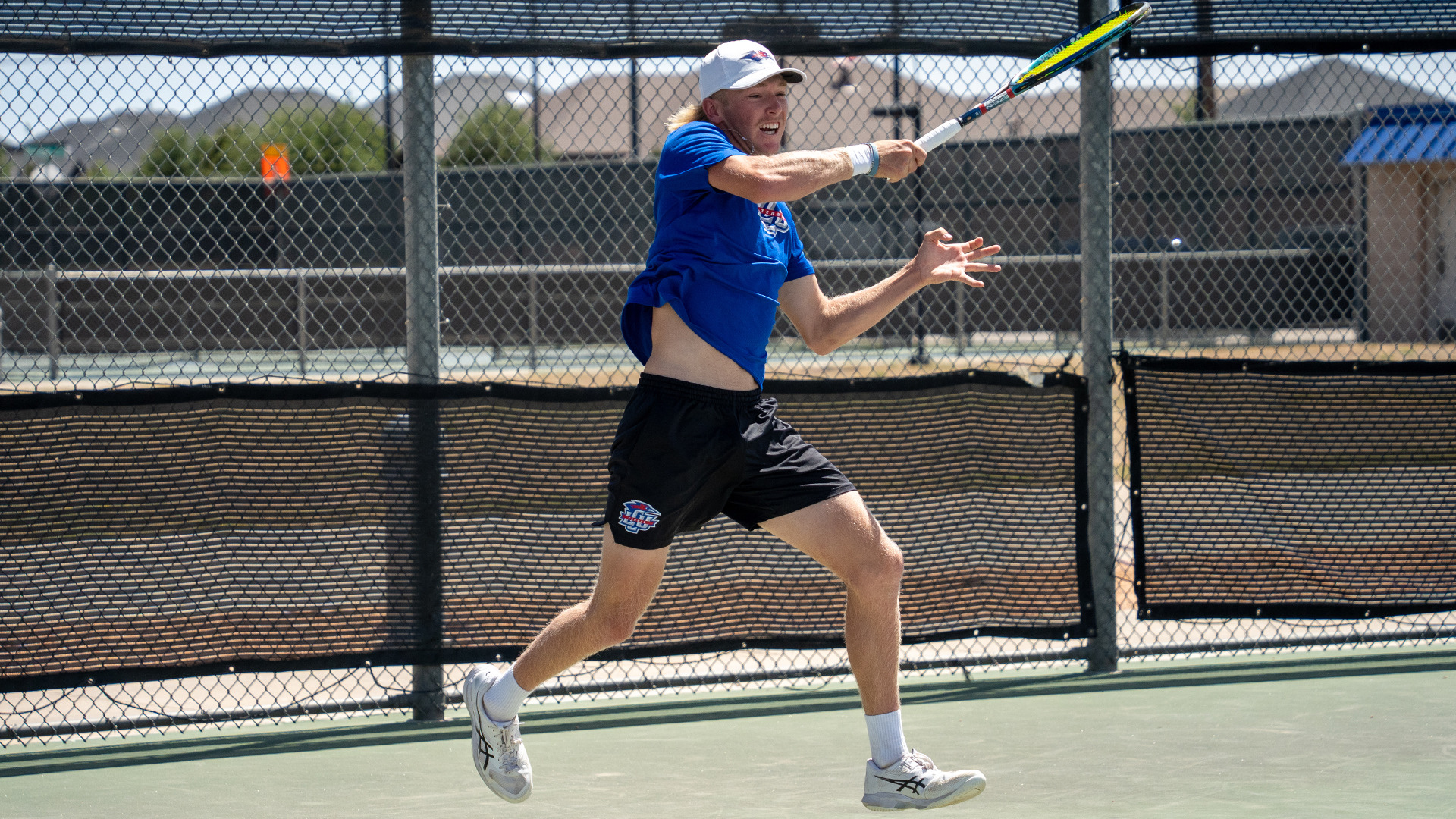 A Chaps men's tennis player in a blue LCU shirt, black shorts, and white cap lunges forward to hit a forehand shot on a sunny outdoor court, with a chain-link fence and buildings visible in the background.